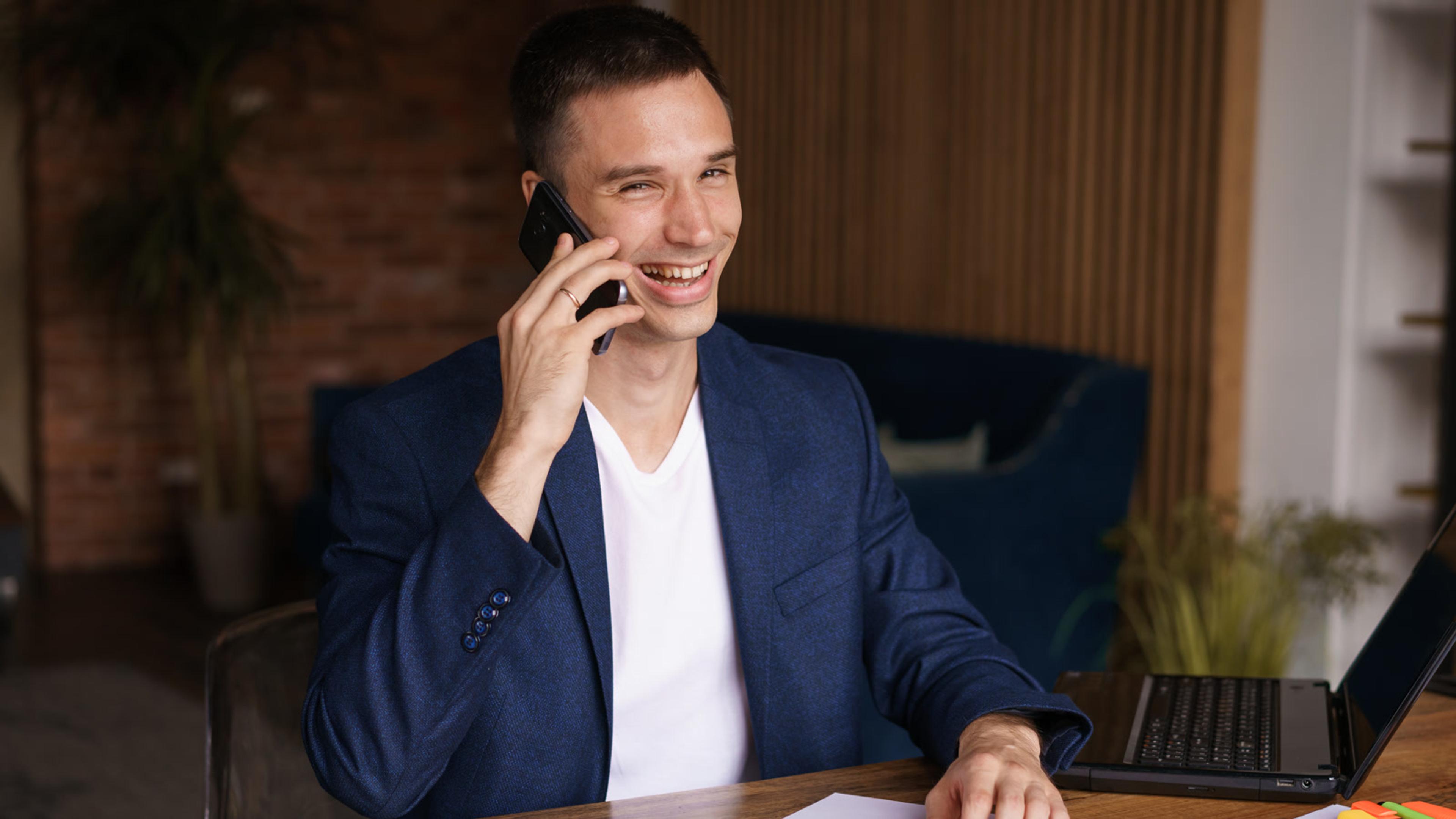 Professional in a blazer smiling while on a smartphone at an office desk with a laptop — BYOD phone under company MDM