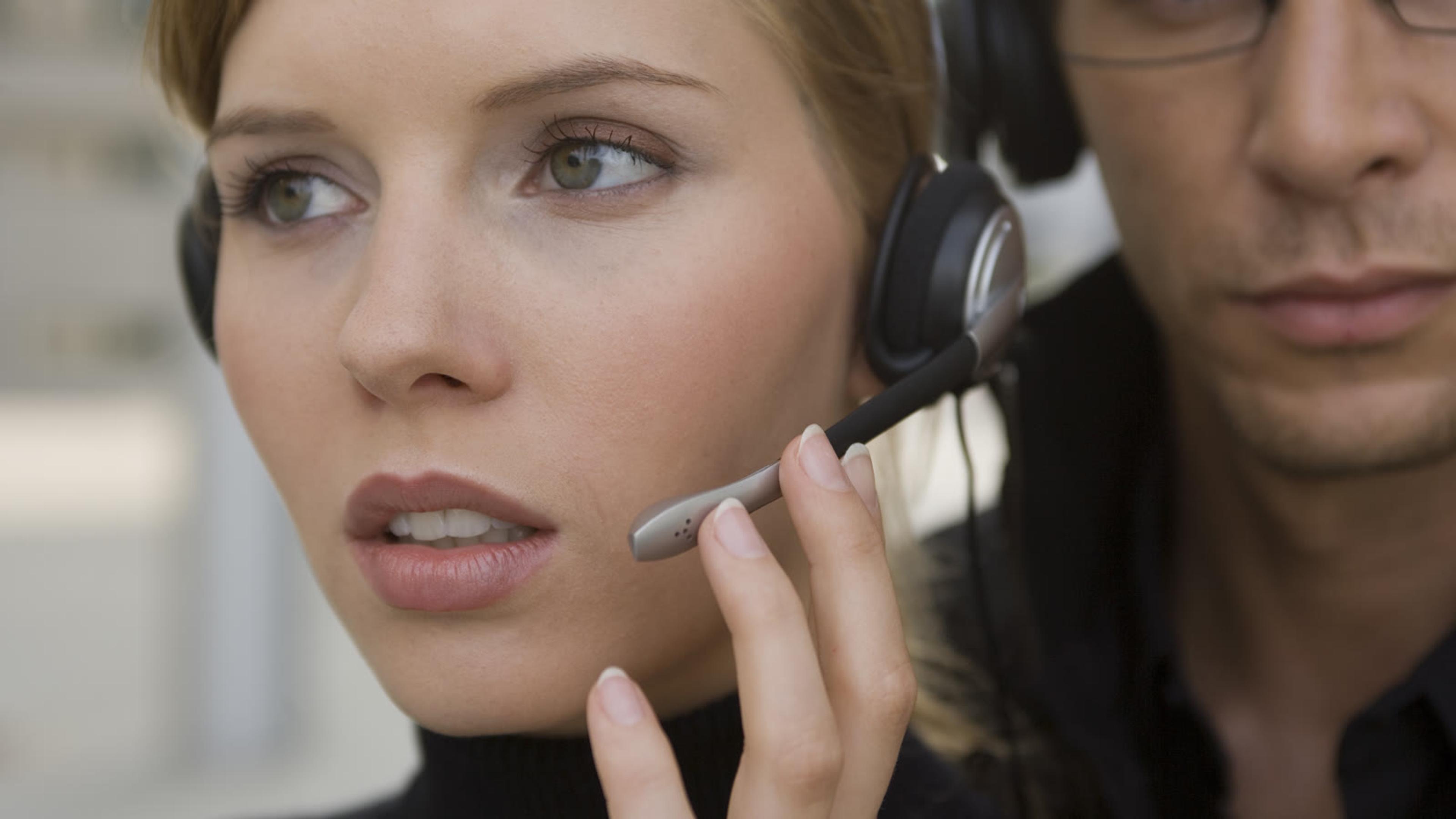 Woman on a headset speaking on a call with a colleague standing behind her