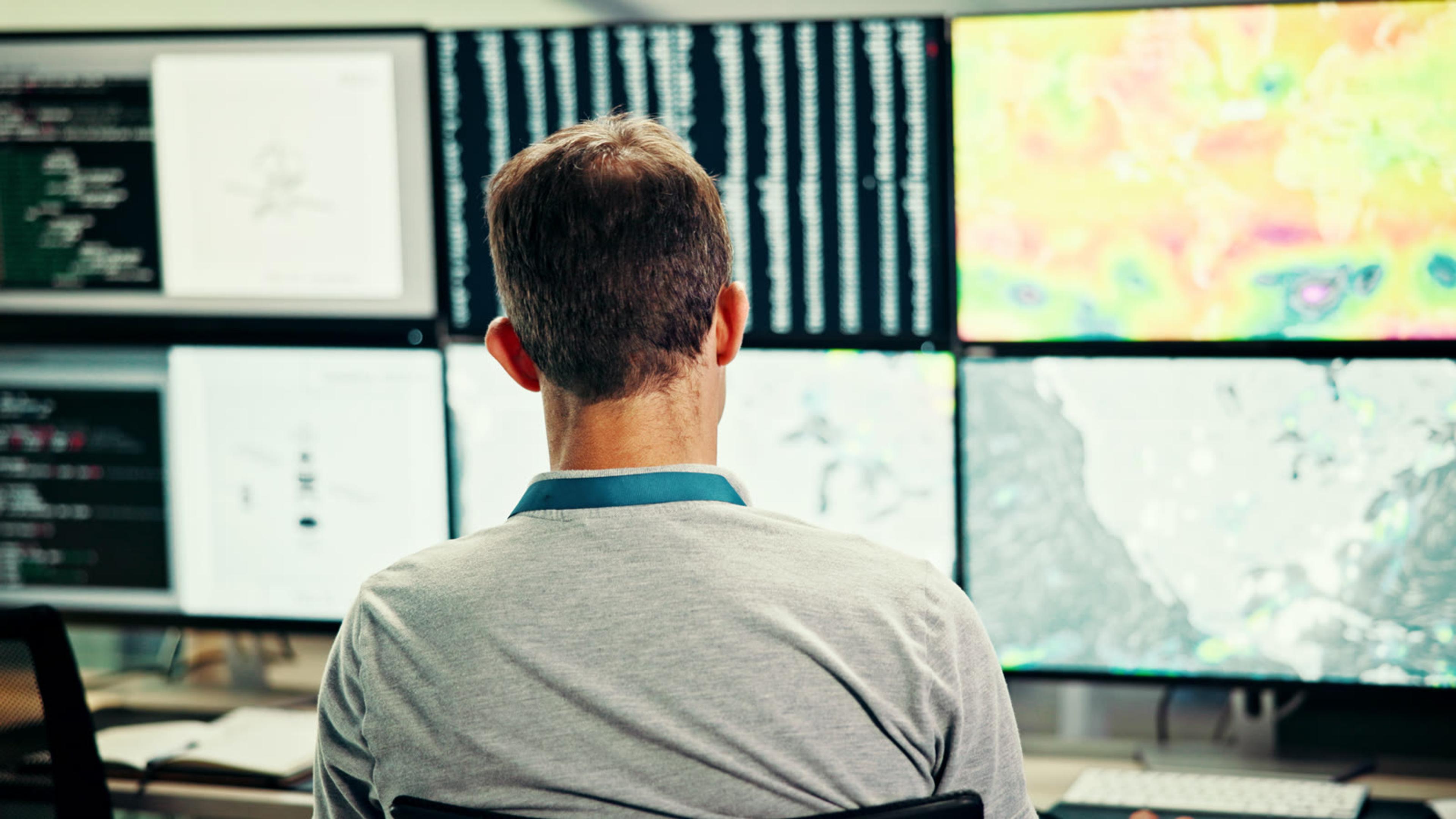 Man seen from behind working at a bank of monitors showing code and map-style data displays
