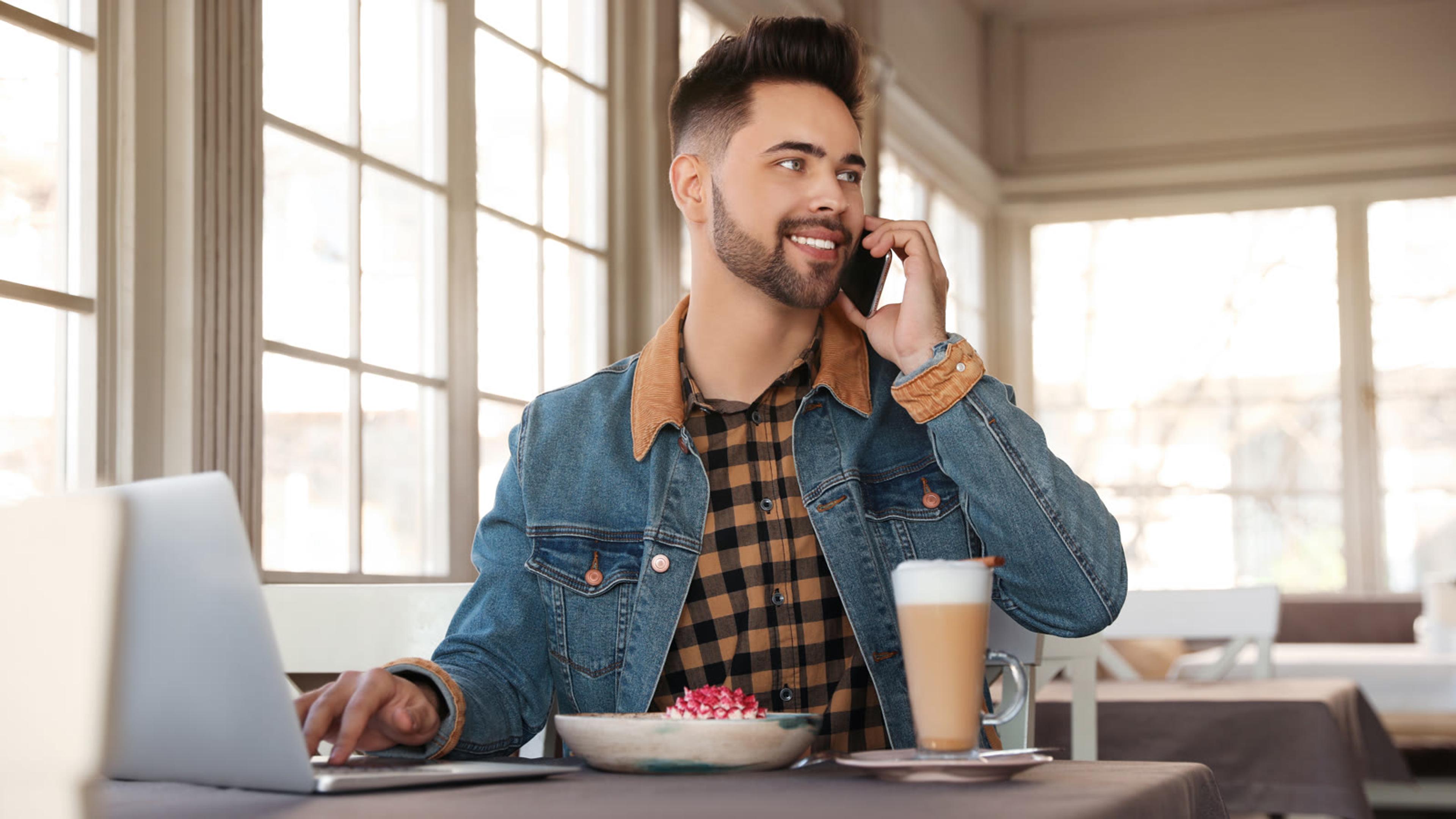 Freelancer taking a business call on his smartphone while working on a laptop in a cafe