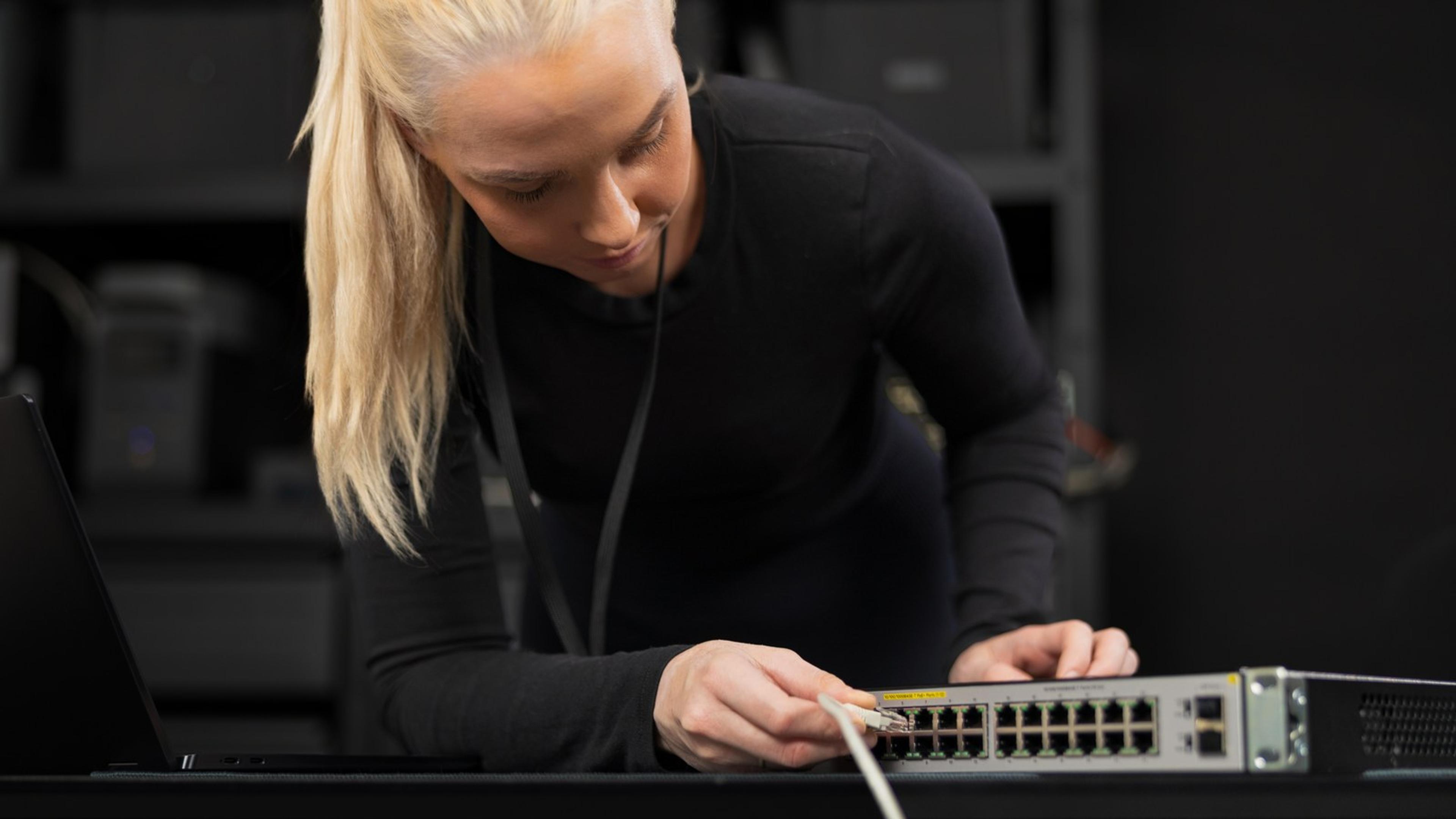 IT admin connecting a cable to a network switch beside an open laptop