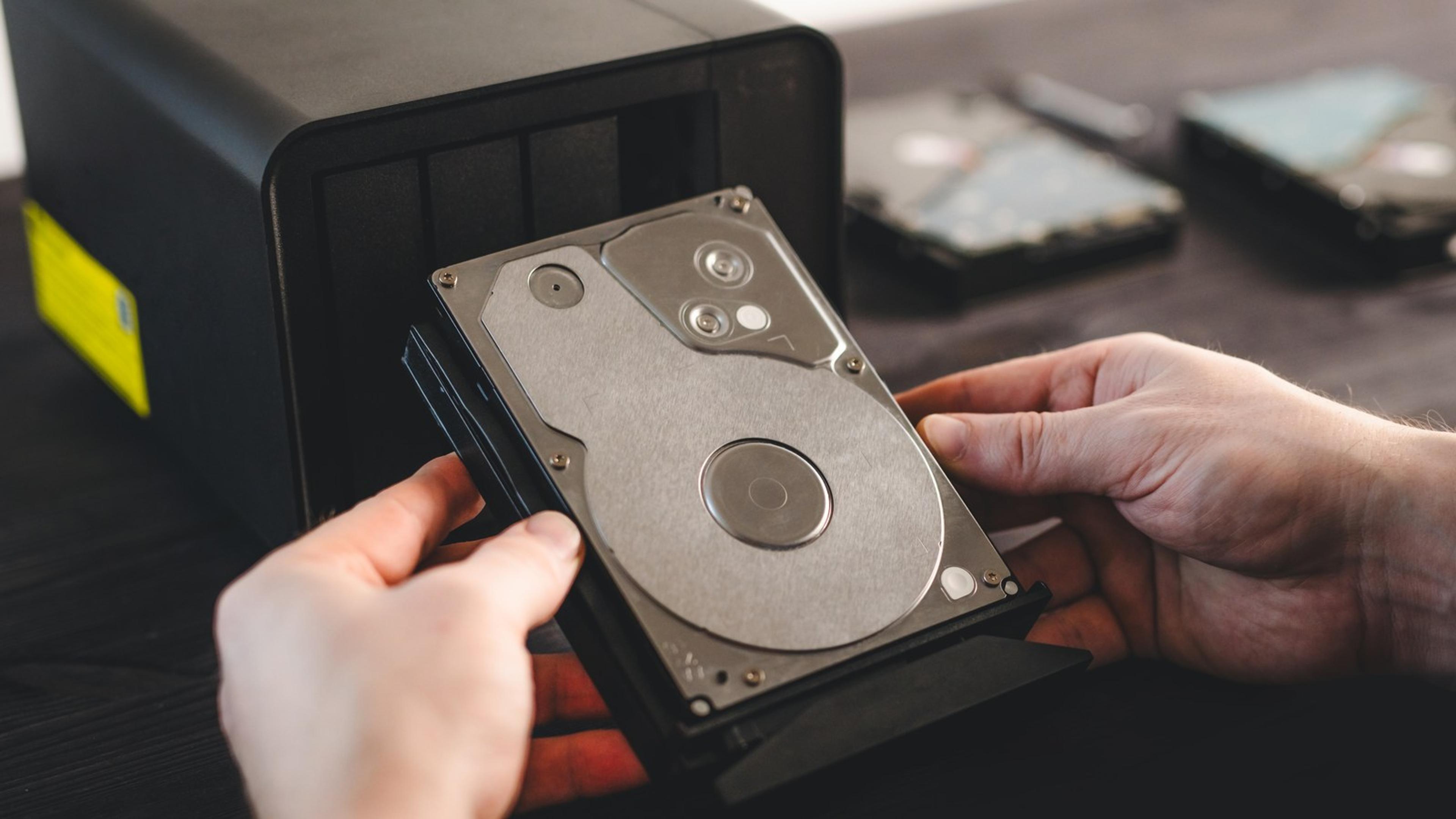 Hands slide a hard drive into a network-attached storage unit on a desk, a backup appliance an attacker cannot delete.