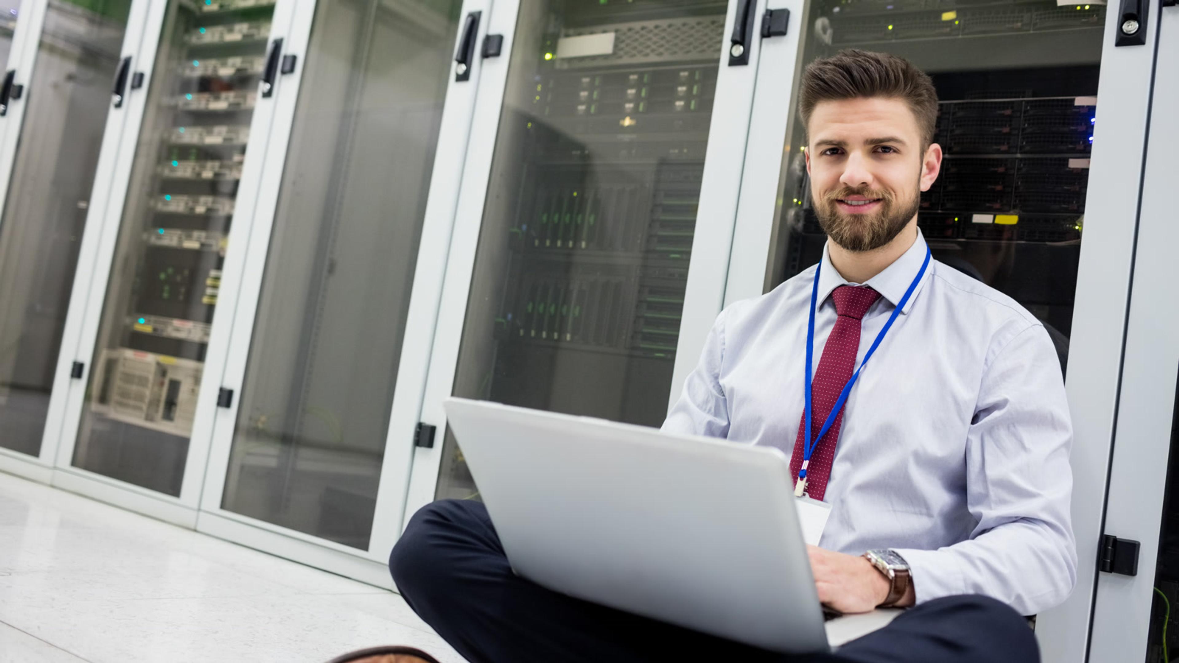 IT professional working on a laptop beside server racks — checking backup jobs and restore readiness