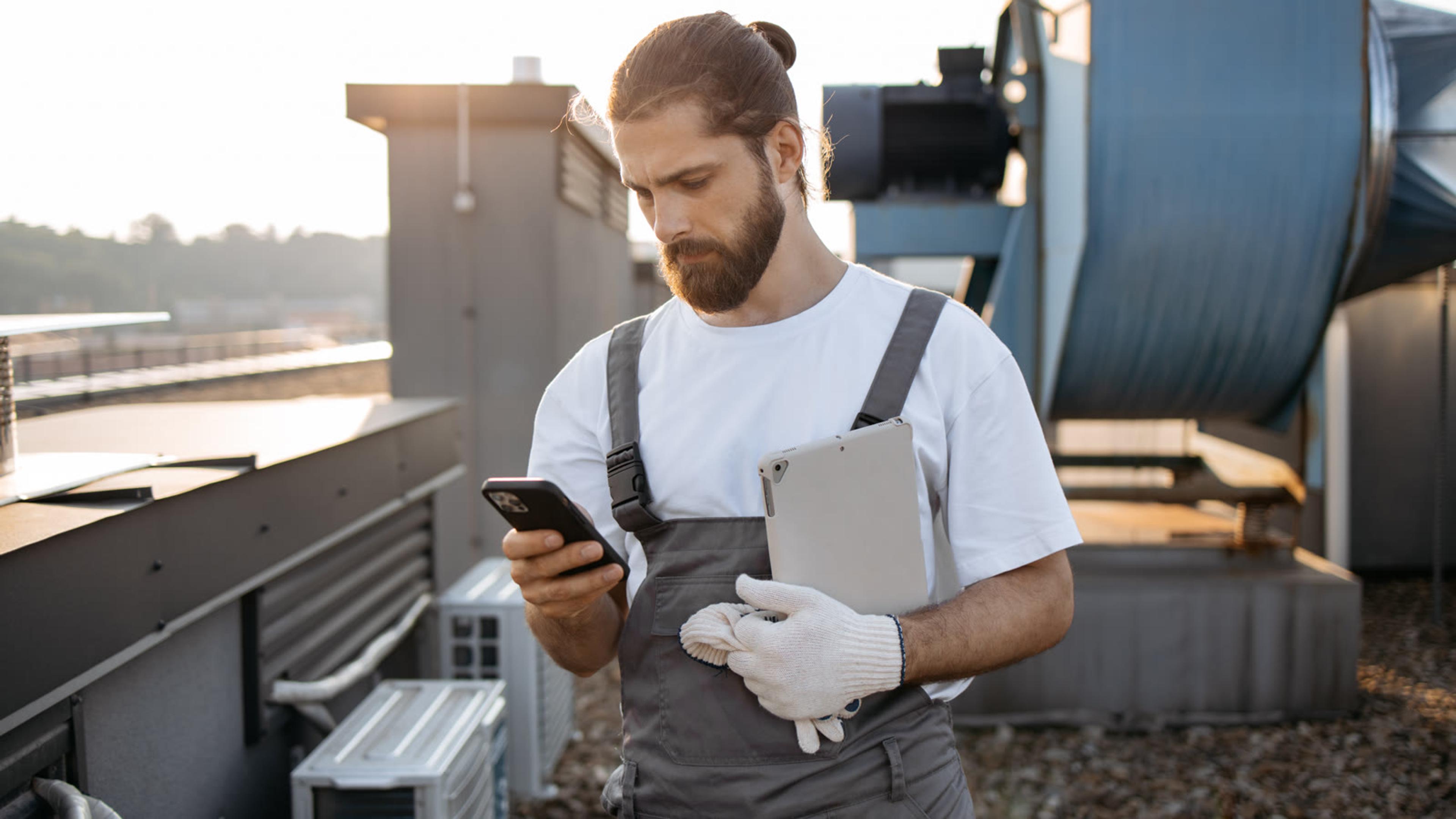 HVAC technician checking a smartphone and tablet on a rooftop — mobile softphone for on-site service