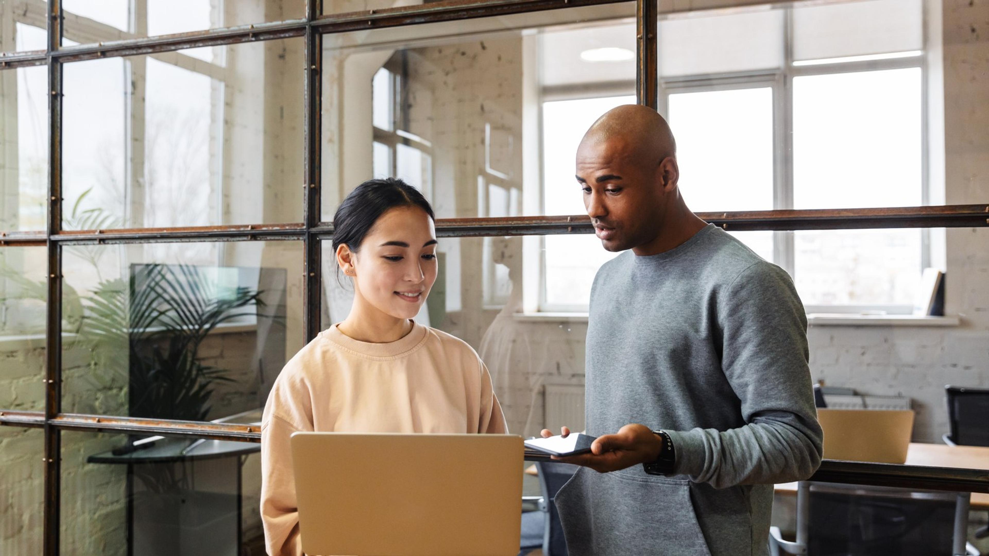 Two colleagues in an office, one holding a laptop and the other a phone