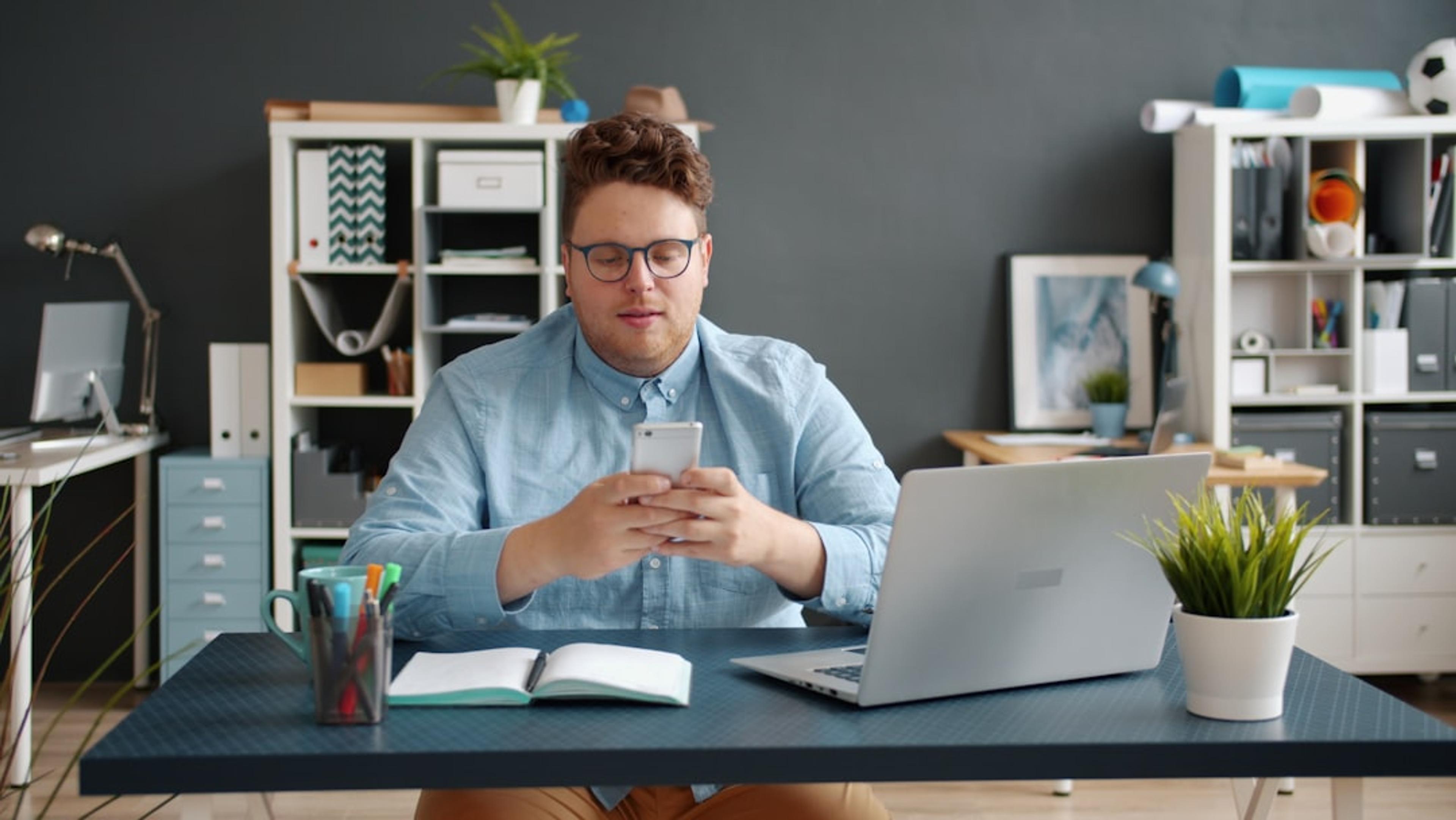 Professional looking at a smartphone at their office desk with a laptop, using a mobile softphone for business communications