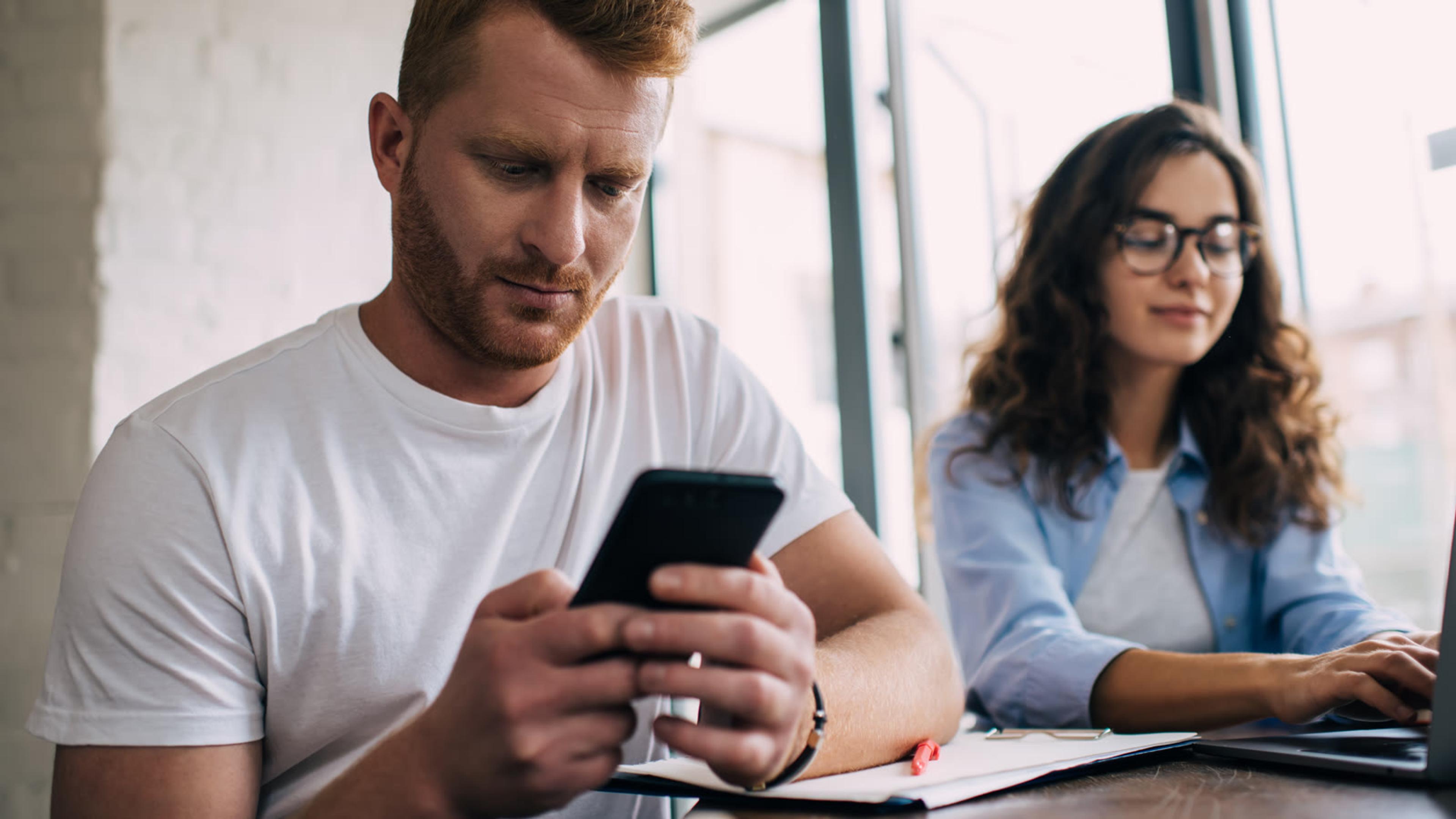 Two colleagues at a table, one checking messages on a smartphone while the other types on a laptop