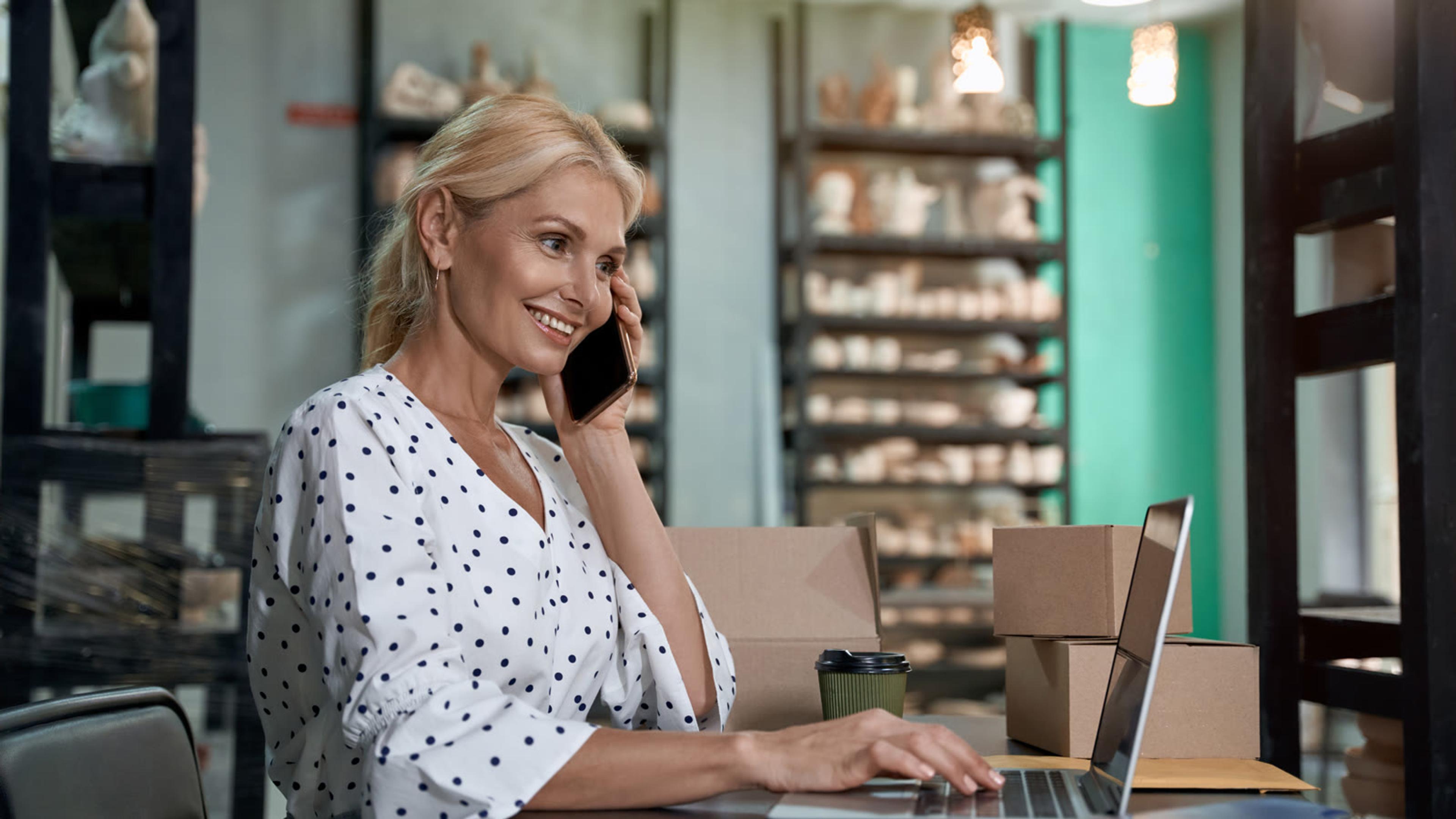 Woman on a smartphone call while working on a laptop at her desk