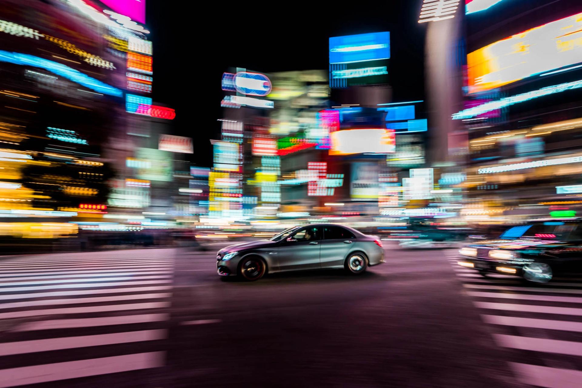 Panning shot of a silver sedan crossing Shibuya at night, the car sharp and the neon city streaked with motion
