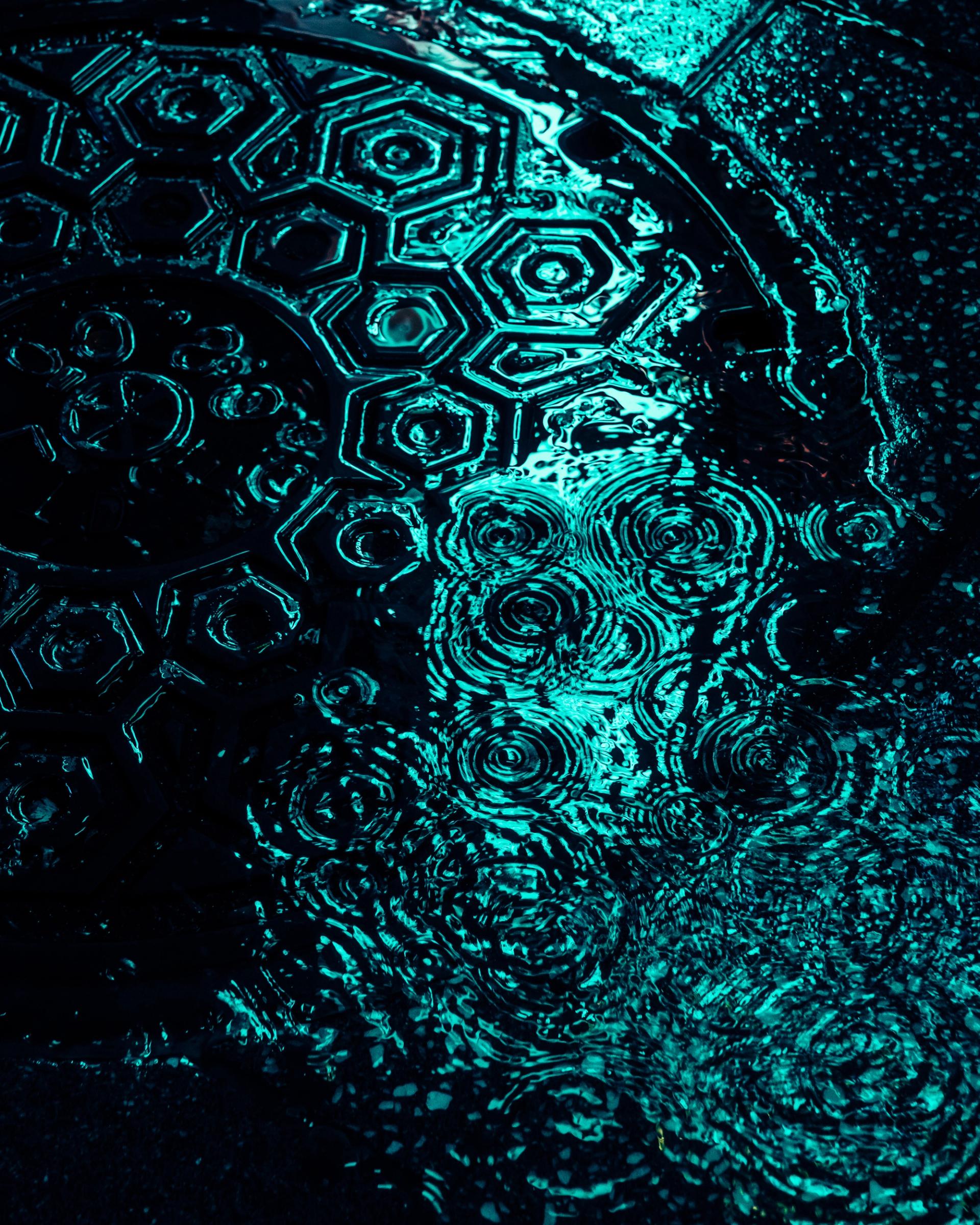 Rain ripples over a manhole cover glowing teal in reflected neon light