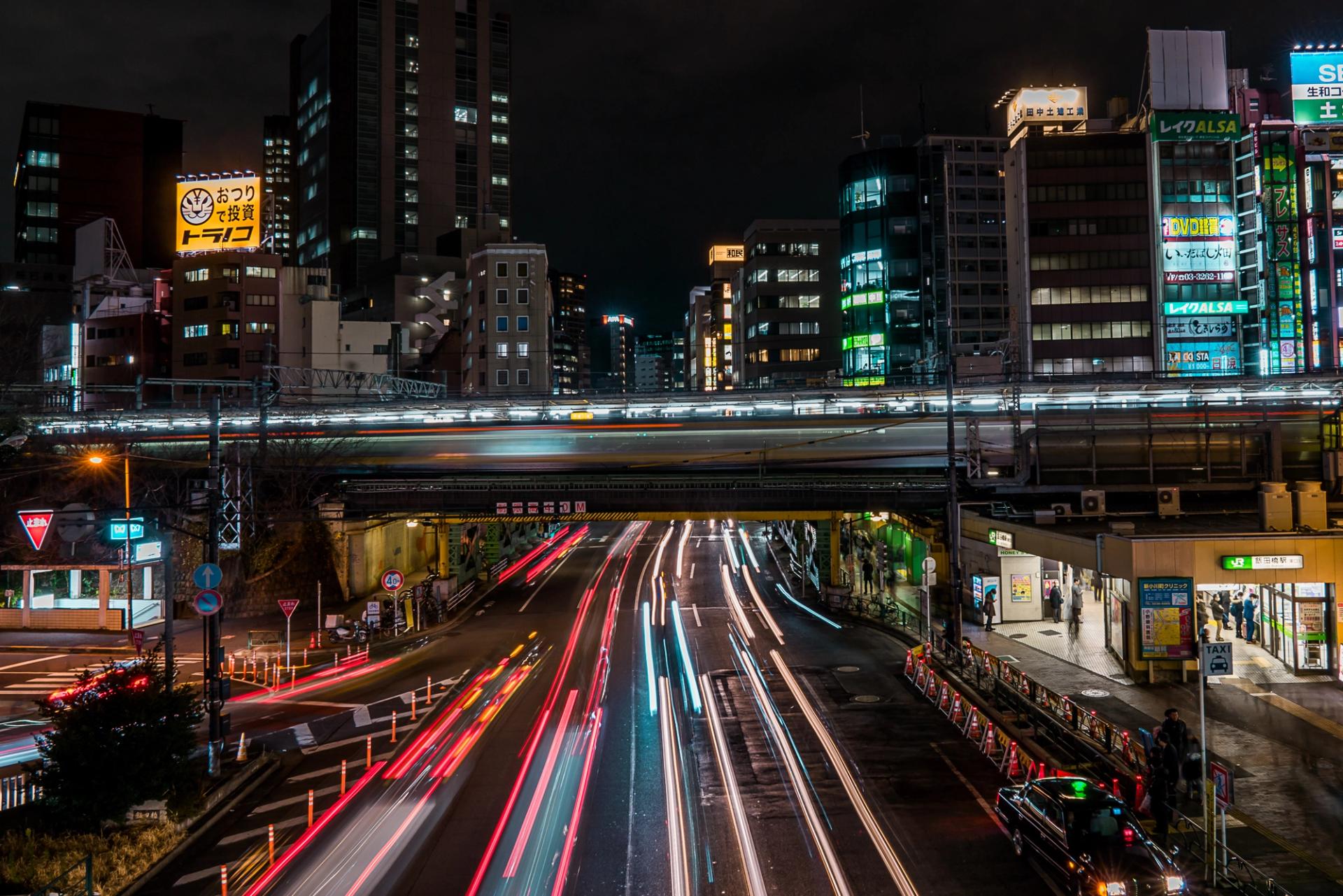 Long exposure over a Tokyo overpass at night, car headlights and taillights drawn into ribbons of light beneath a streaking train