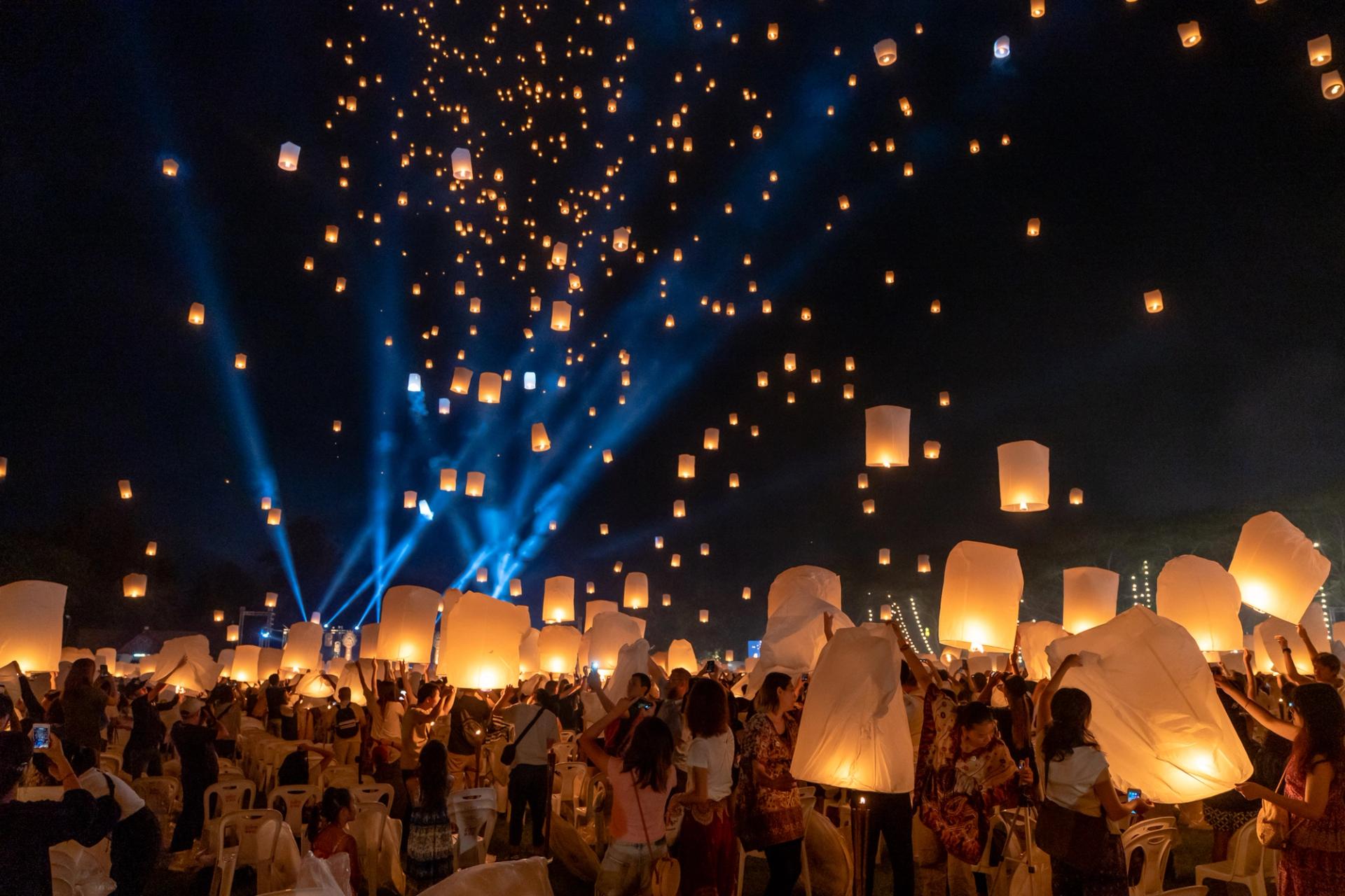 Sky lantern festival at a fast shutter, hundreds of lanterns held still in the air above the crowd with blue stage beams behind