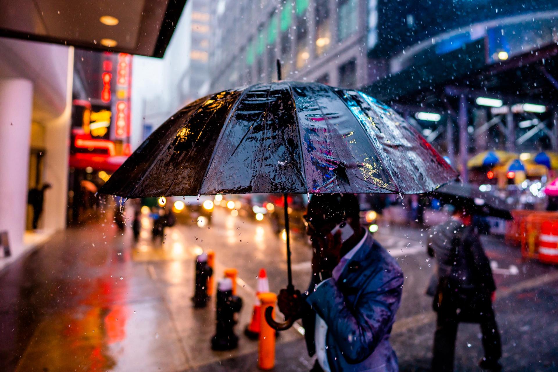 Man under a black umbrella in a downpour, raindrops frozen in the air against neon signs and traffic lights