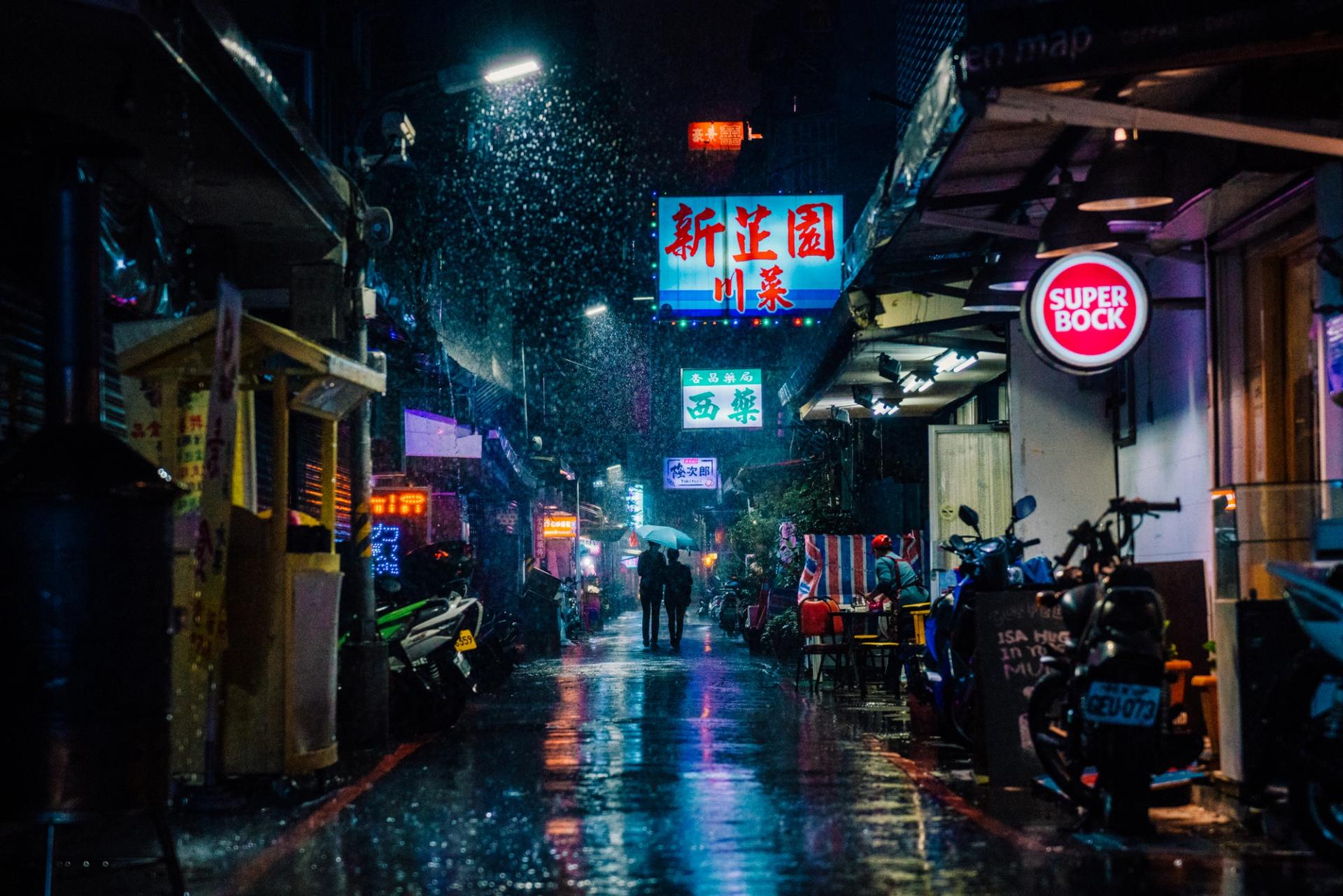 Rain frozen in the light beams of a neon-lit alley at night, a couple walking under an umbrella in the distance