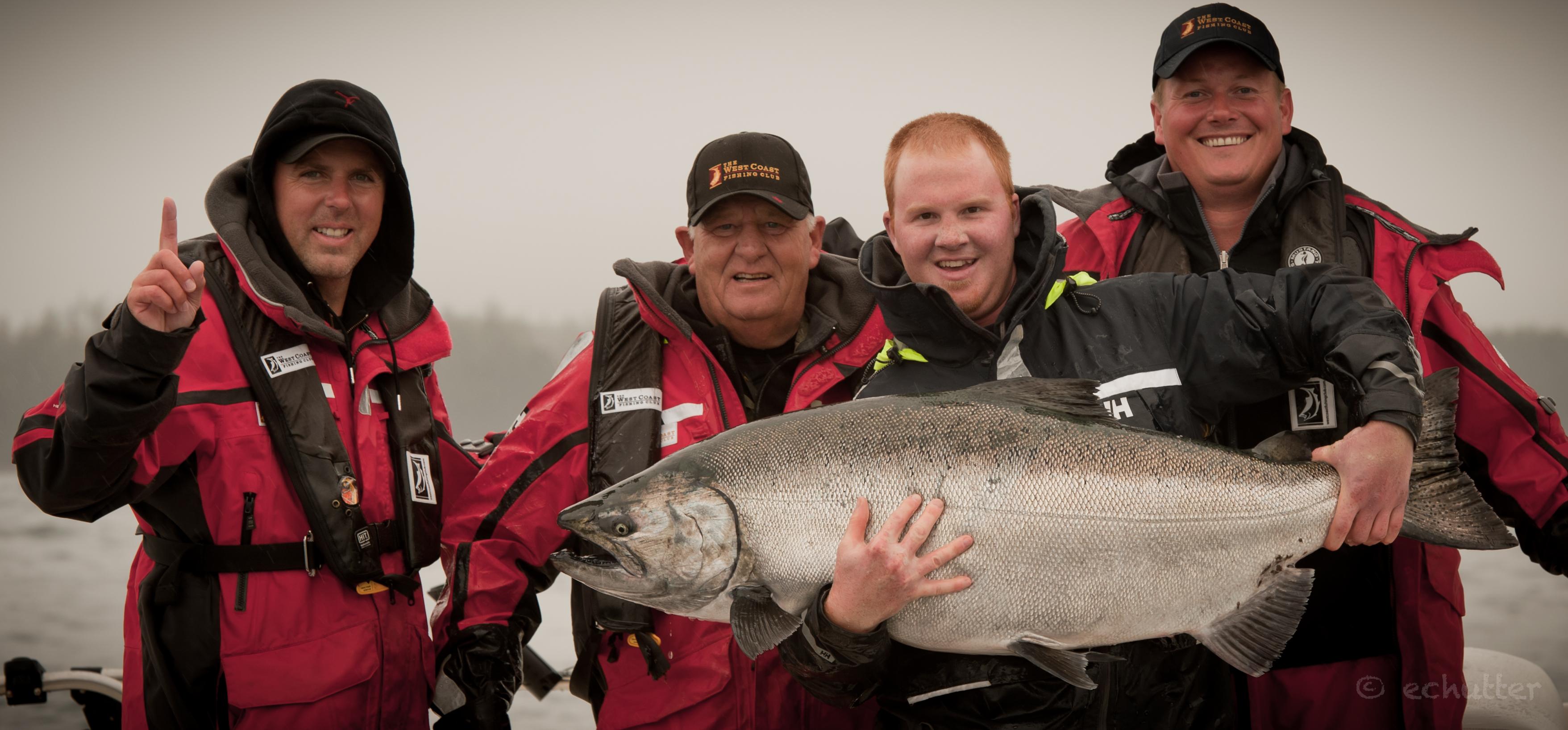 Four anglers in survival suits holding up a large salmon on the dock