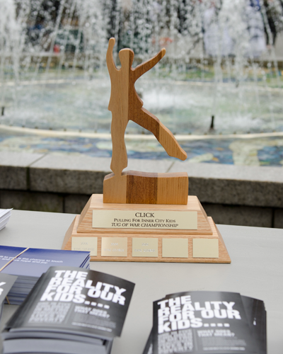 The wooden Pulling for Inner City Kids tug of war championship trophy on a table beside stacks of CLICK T-shirts