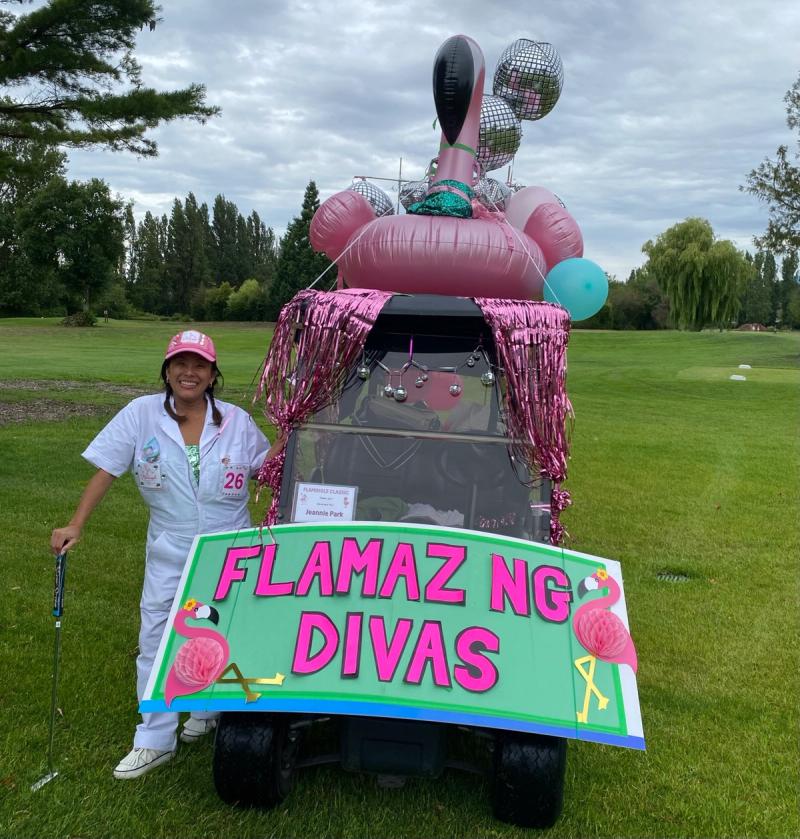 A golfer in white overalls stands beside a golf cart decorated with an inflatable flamingo, disco balls, pink tinsel and a sign reading Flamazing Divas.