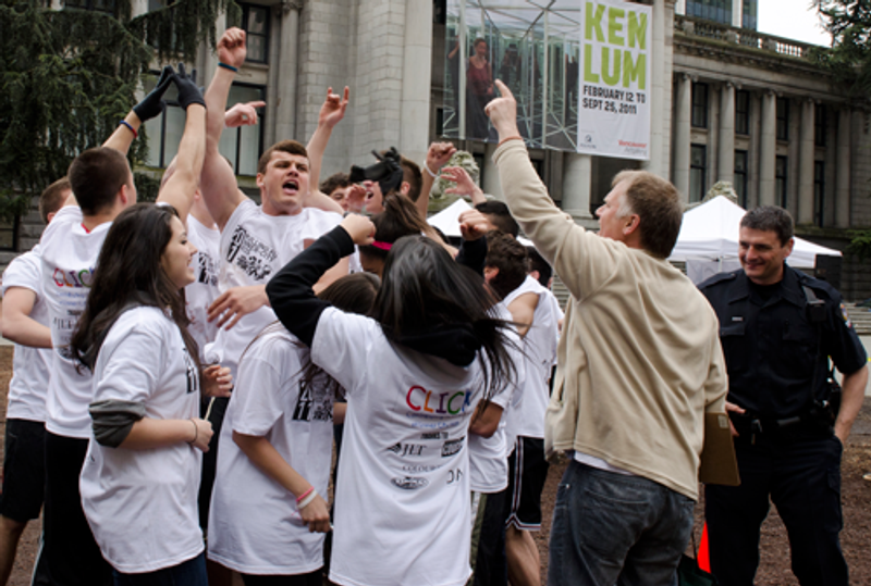 Students in white CLICK T-shirts throw their arms up in victory beside a police officer