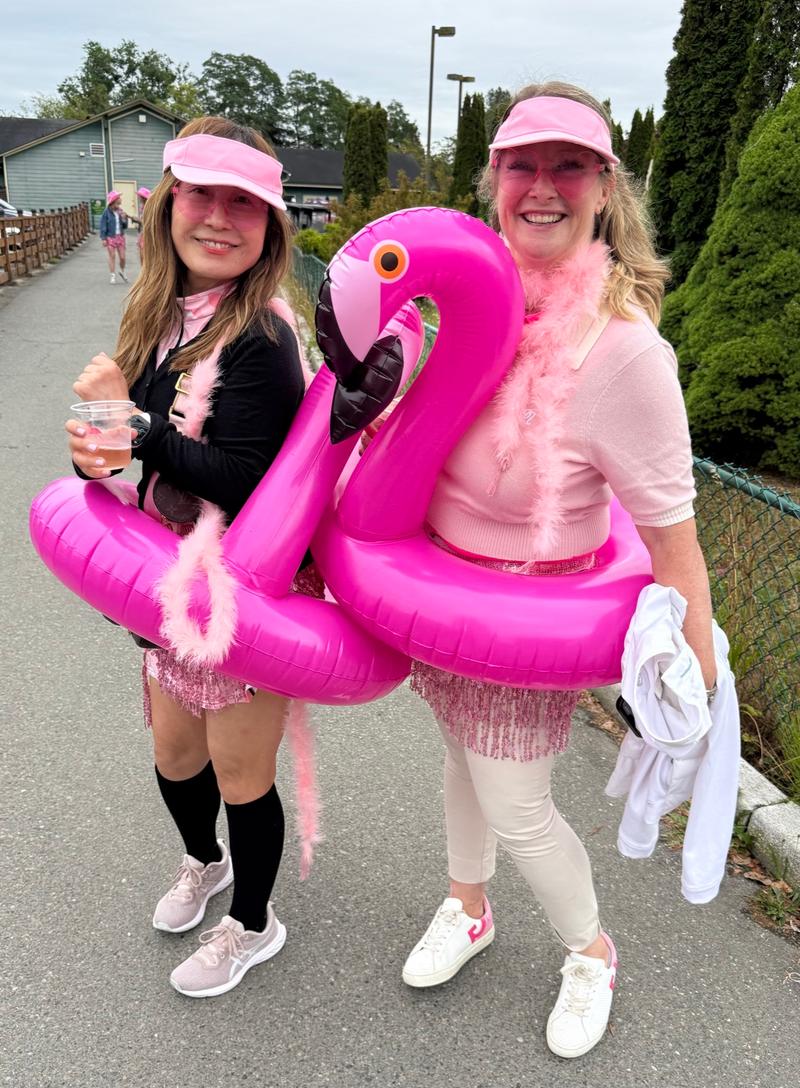 Two players in pink visors and feather boas wear inflatable pink flamingo rings on the path beside the course.