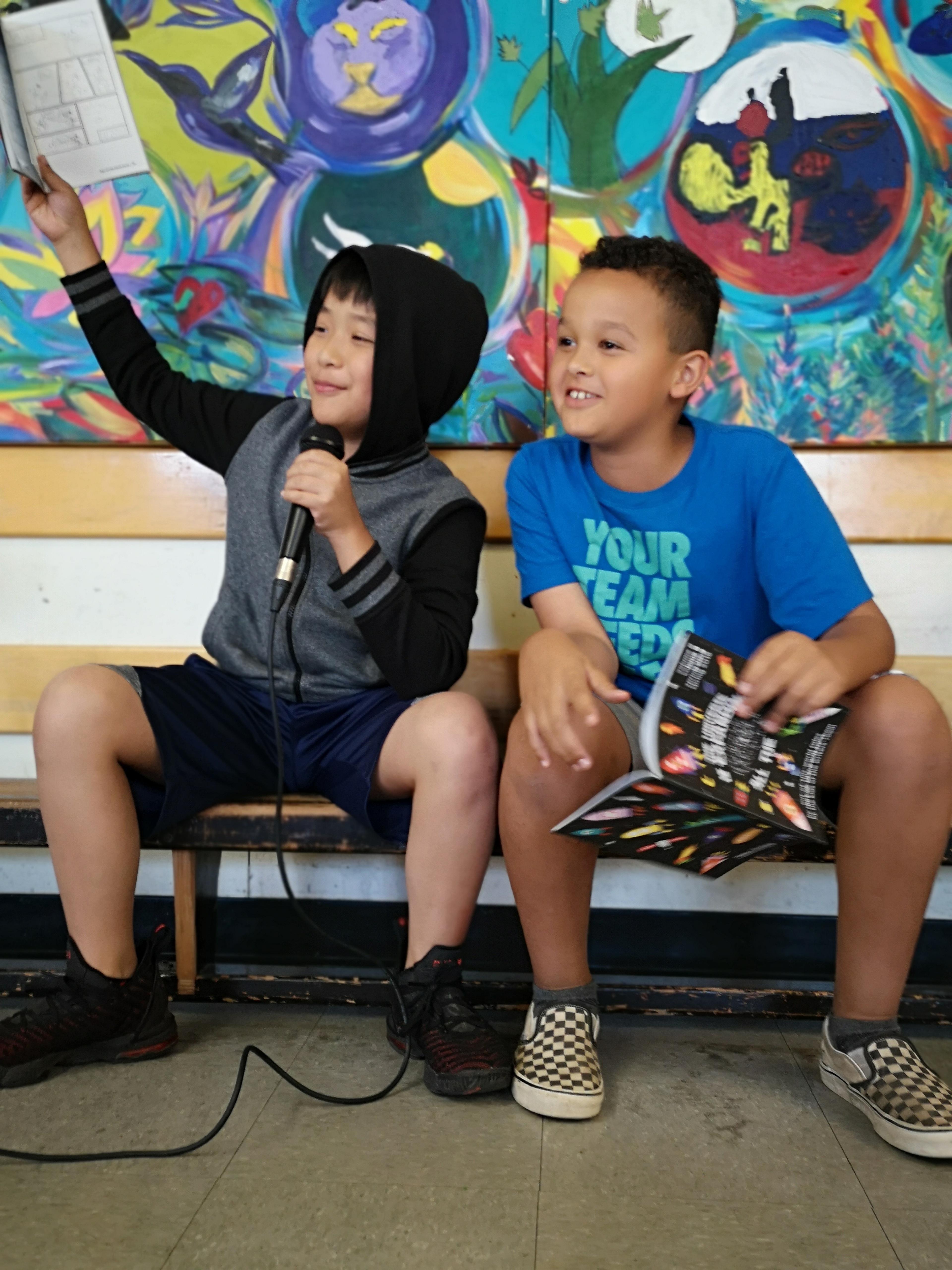 Two children at The Writers' Exchange, one holding up a page and speaking into a microphone in front of a painted mural