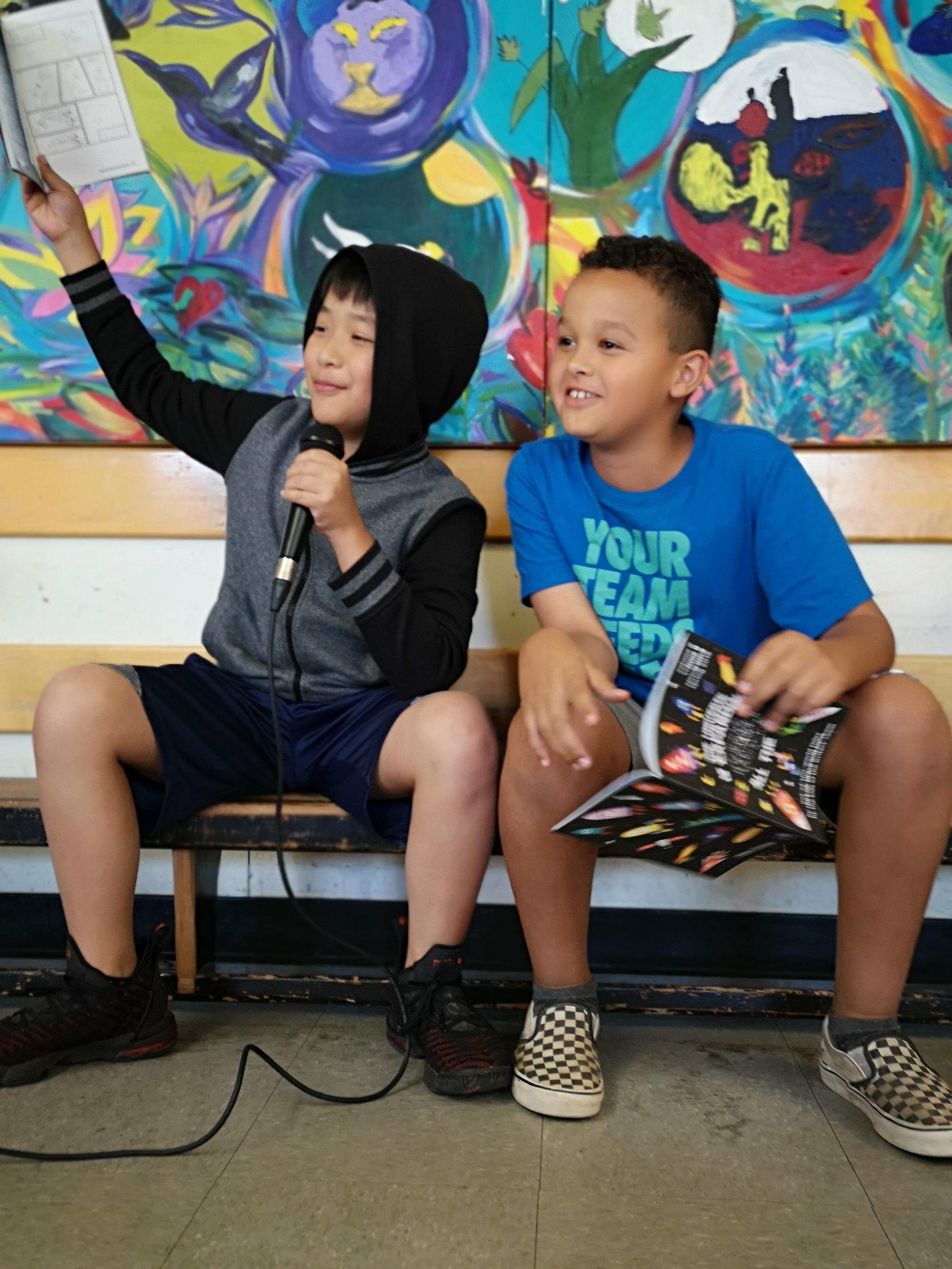 Two children at The Writers' Exchange, one holding up a page and speaking into a microphone in front of a painted mural