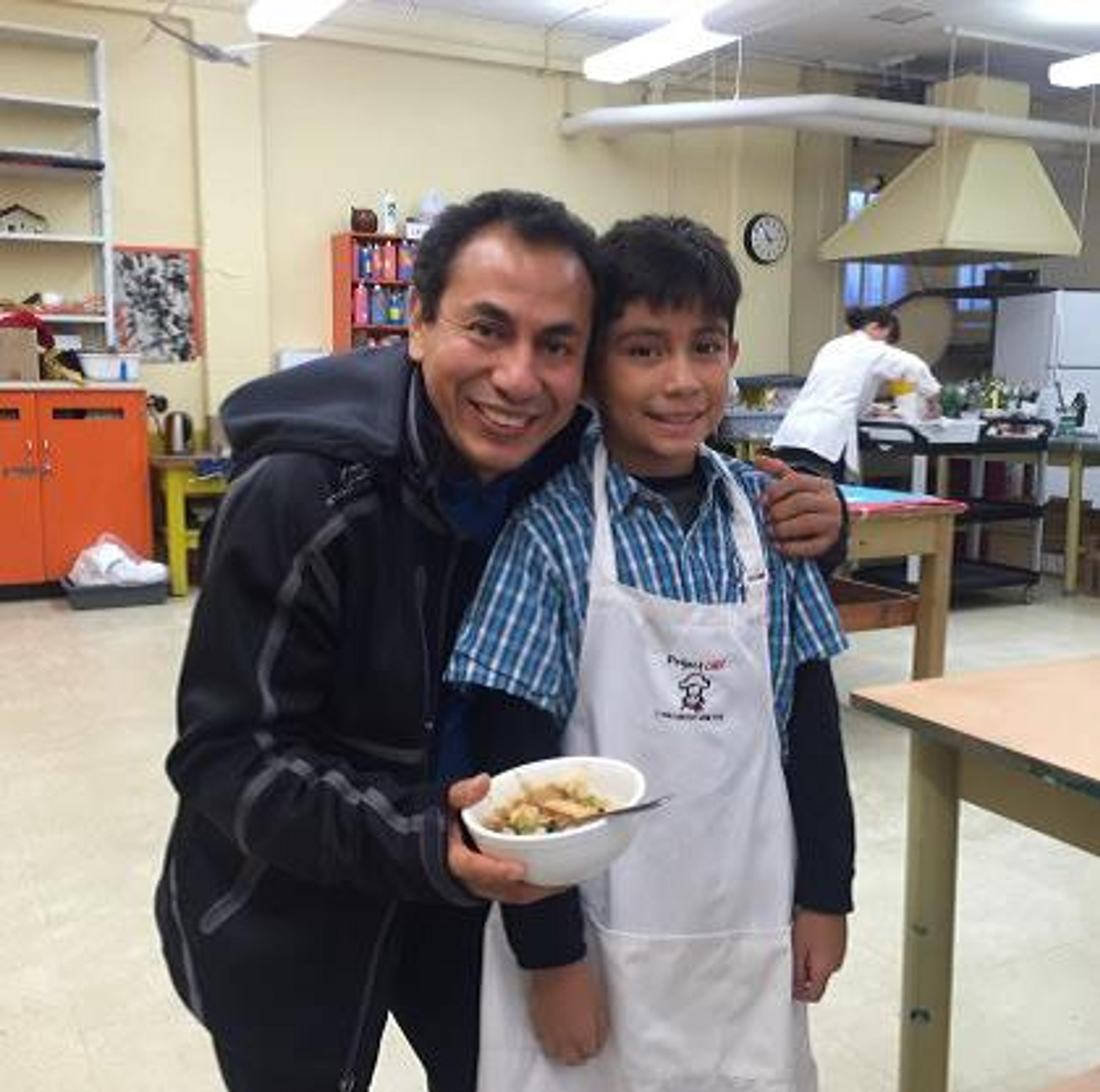 A Project CHEF instructor and a student in aprons in a school kitchen, holding a bowl of food they made