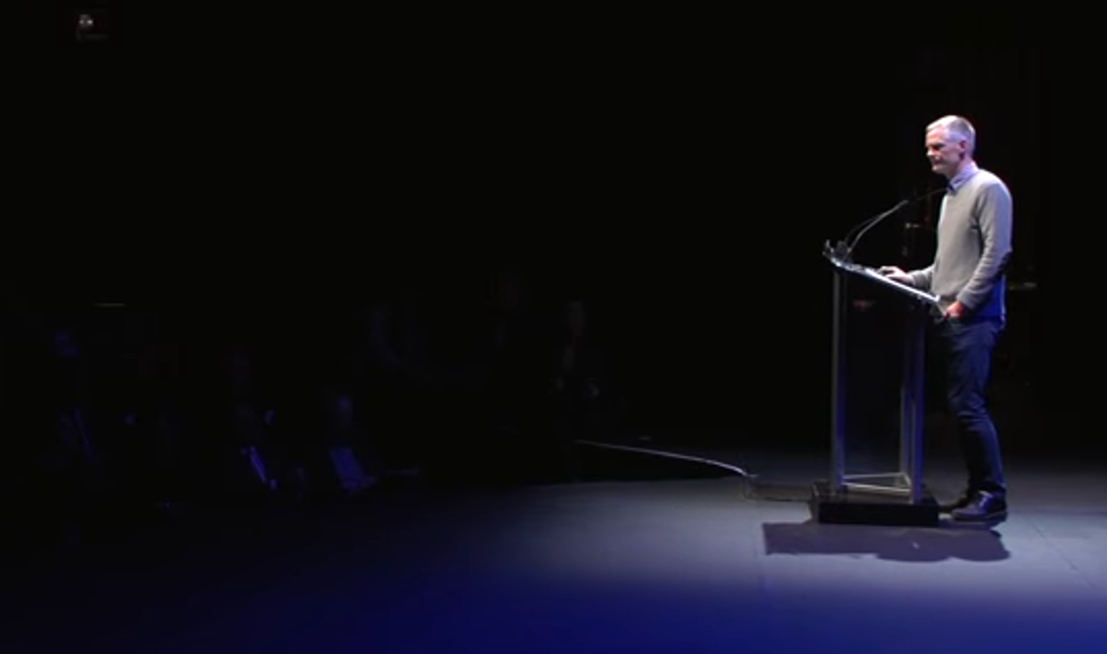 Trevor Stokes speaking at a podium under stage lighting at Sam Sullivan's Public Salon