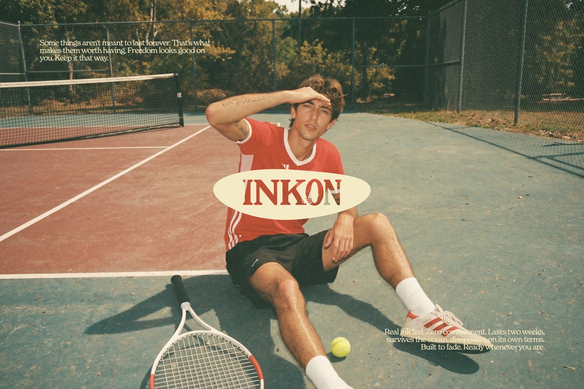 A man in a red shirt sitting on a tennis court beside a racket, under the INKON logo