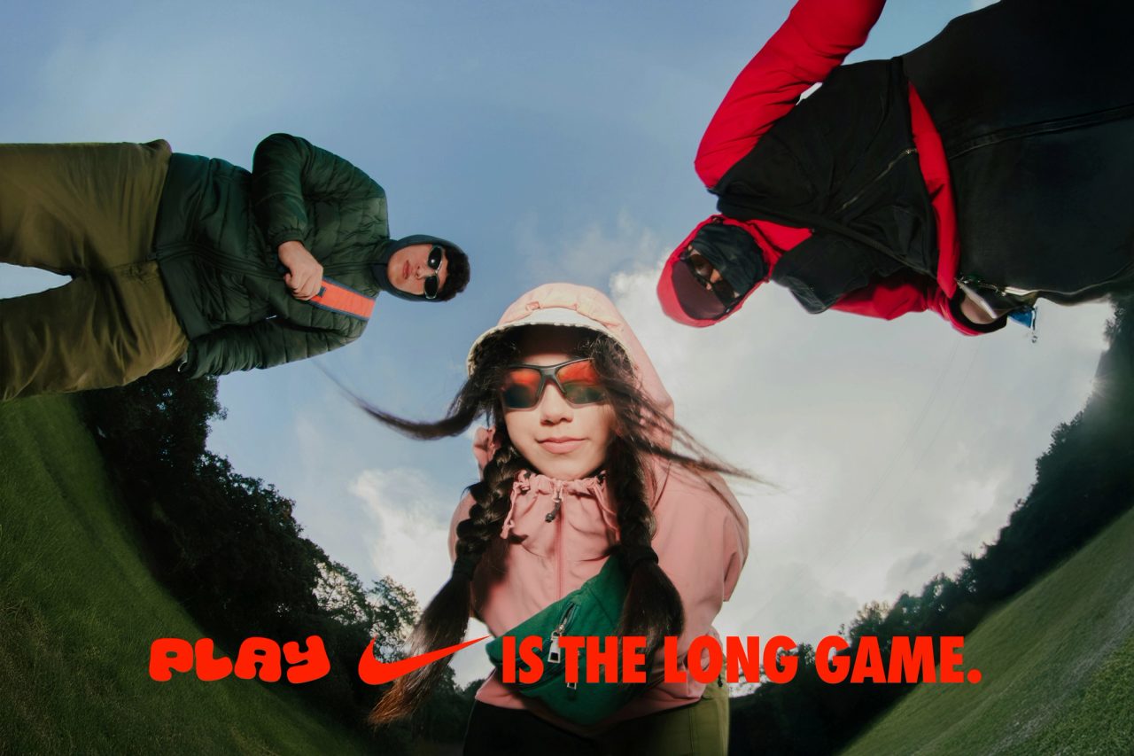 Three friends in outdoor gear peer into a fisheye lens, captioned Play Is the Long Game