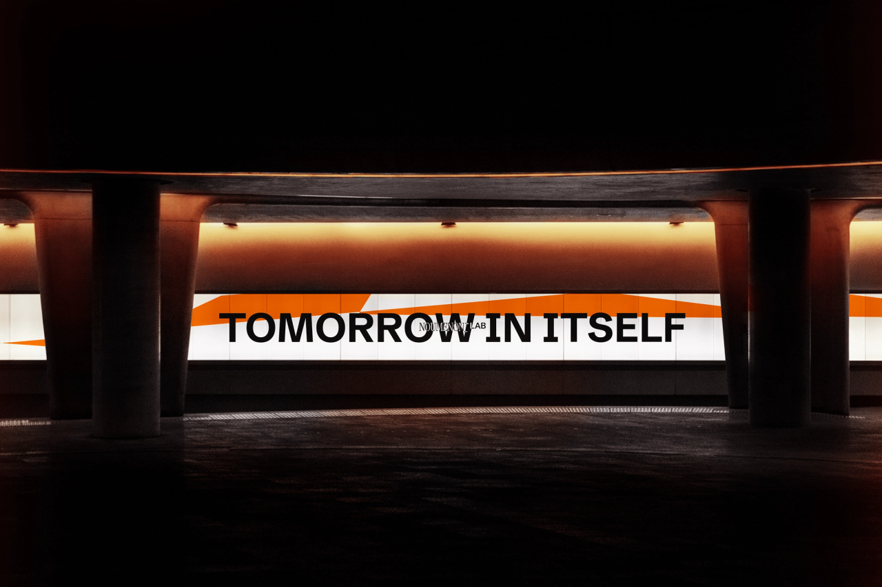 Backlit sign reading "Tomorrow in itself" with the NOUMENON LAB logo, between concrete pillars
