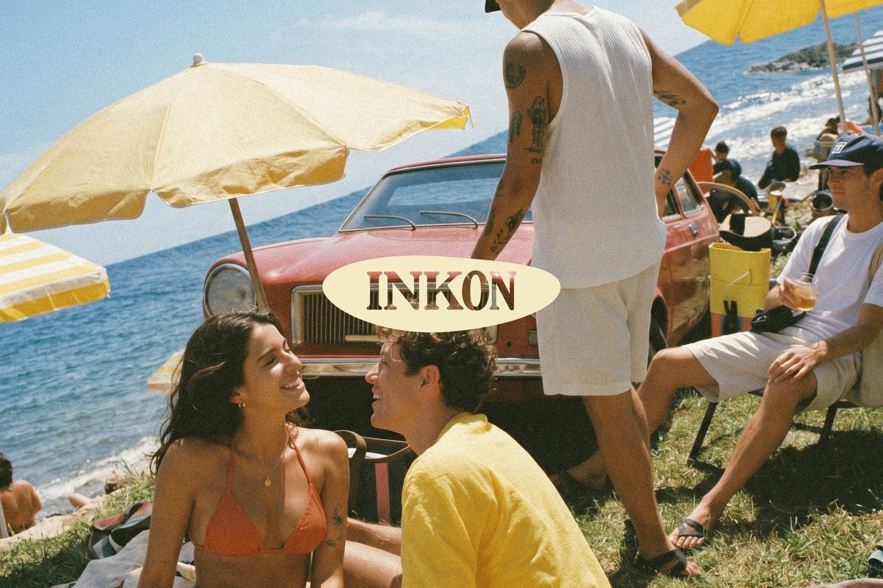 Beachgoers by a red vintage car and yellow umbrellas behind the INKON oval logo