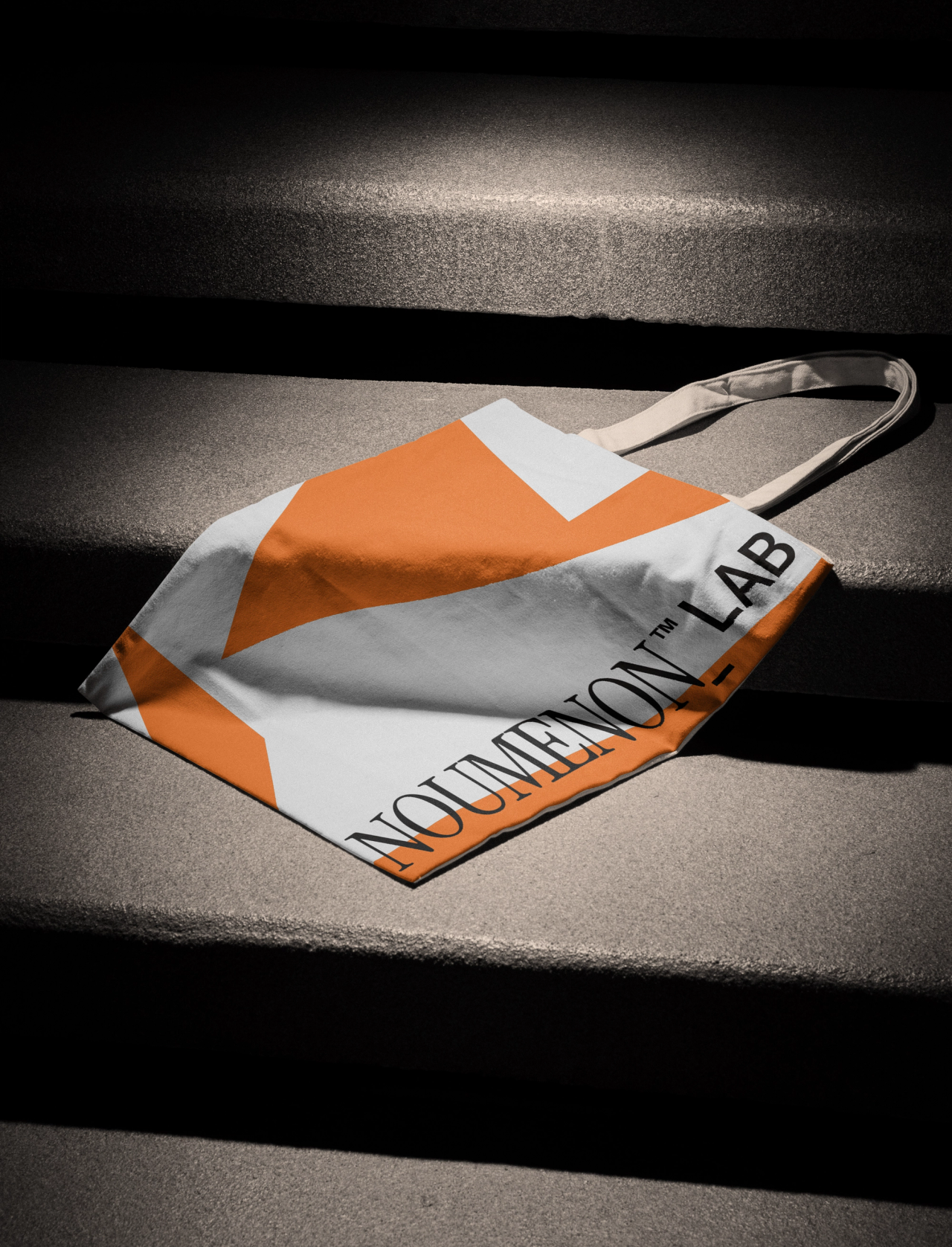 An orange and white NOUMENON LAB tote bag on concrete steps in a shaft of light