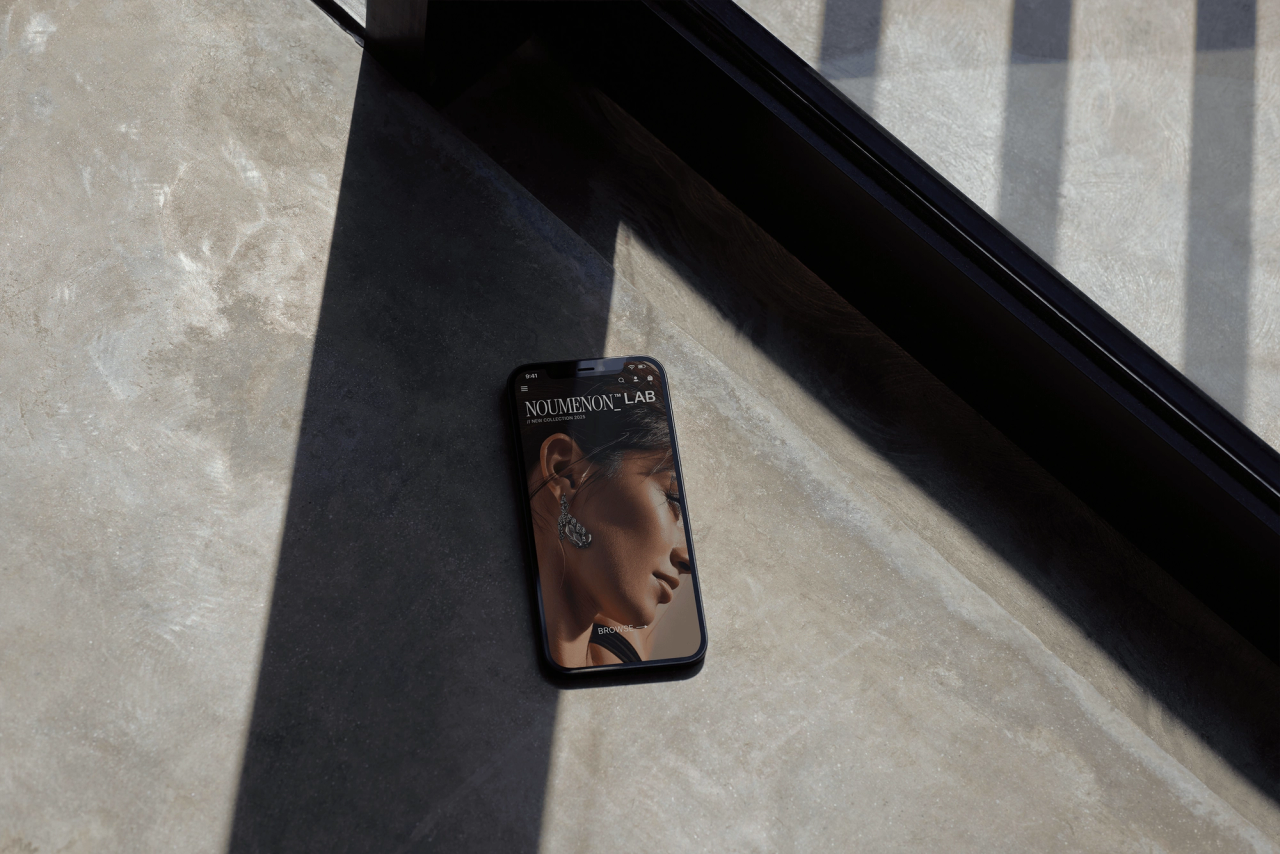 A phone on sunlit concrete showing a woman's silver earring on a NOUMENON LAB page