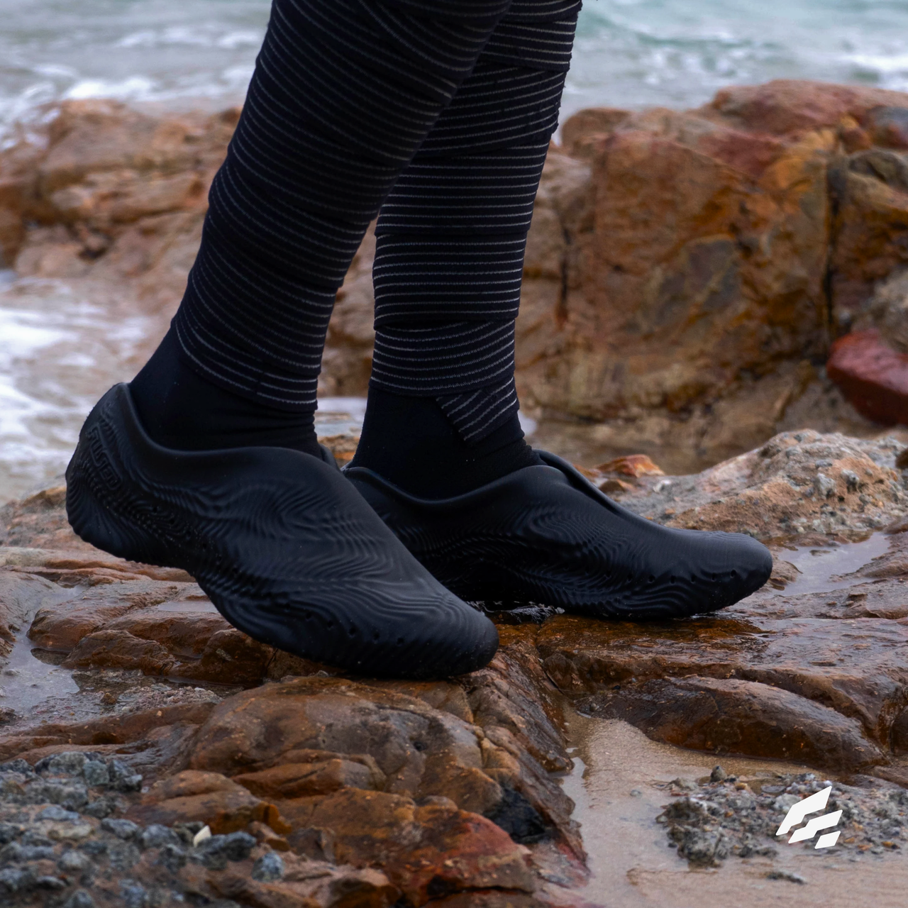 A pair of black 3D-printed shoes worn on wet seaside rocks, with the Fused mark