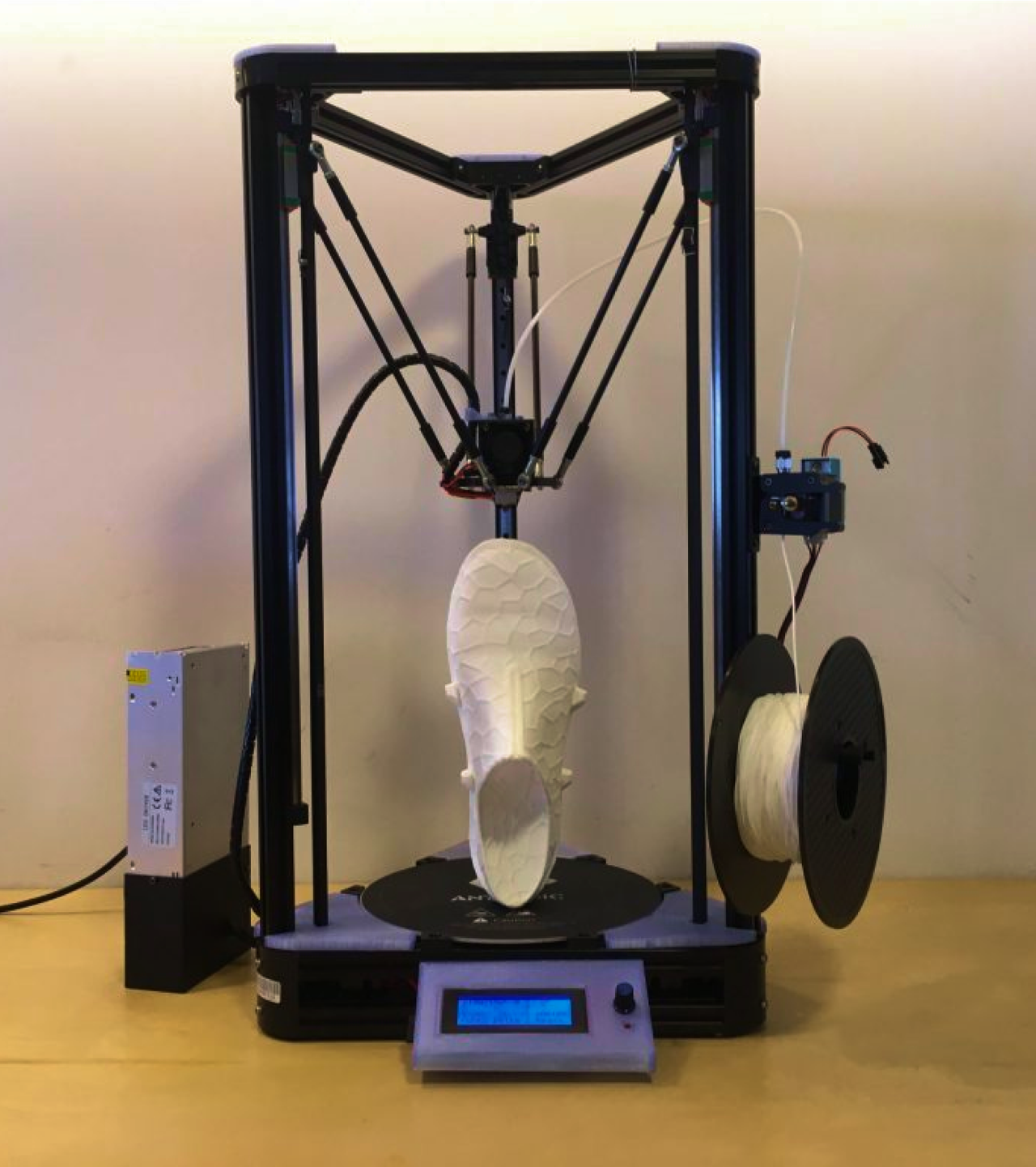 A 3D printer with a white shoe on its build plate beside a filament spool