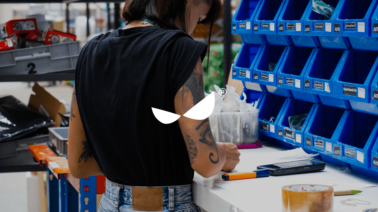 A tattooed worker at a workbench by blue storage bins, behind the Casa de Héroes symbol