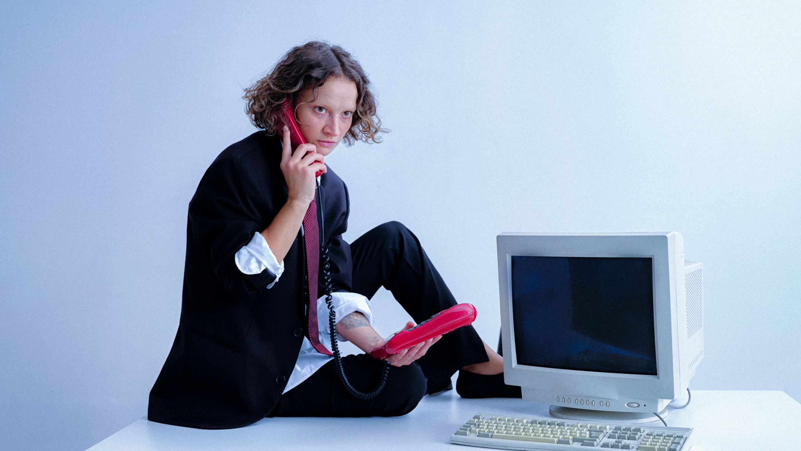 A woman in a black suit on an office desk, holding a red phone