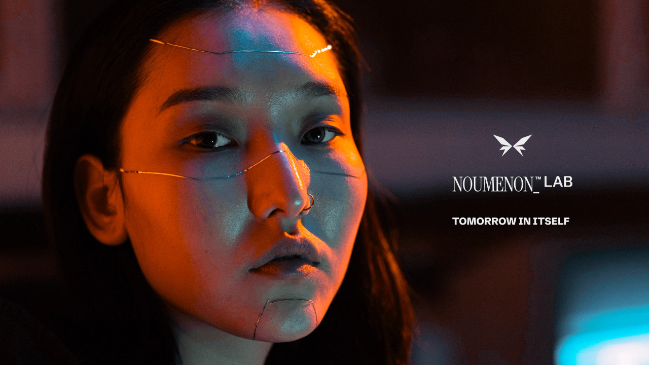 A woman with thin wires across her face in orange light, beside the NOUMENON LAB logo