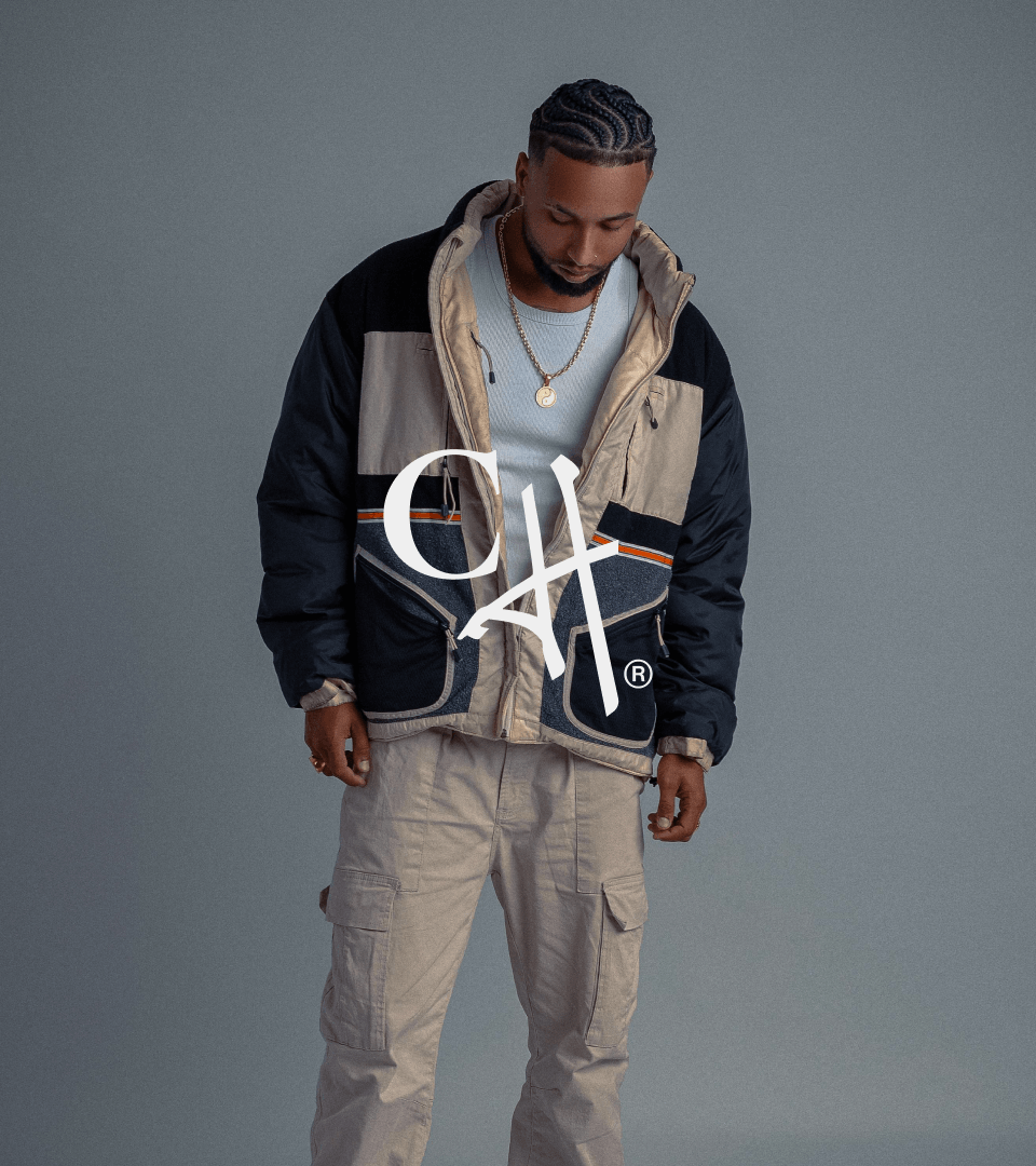 A man in a patchwork puffer jacket and cargo trousers behind the Casa de Héroes monogram