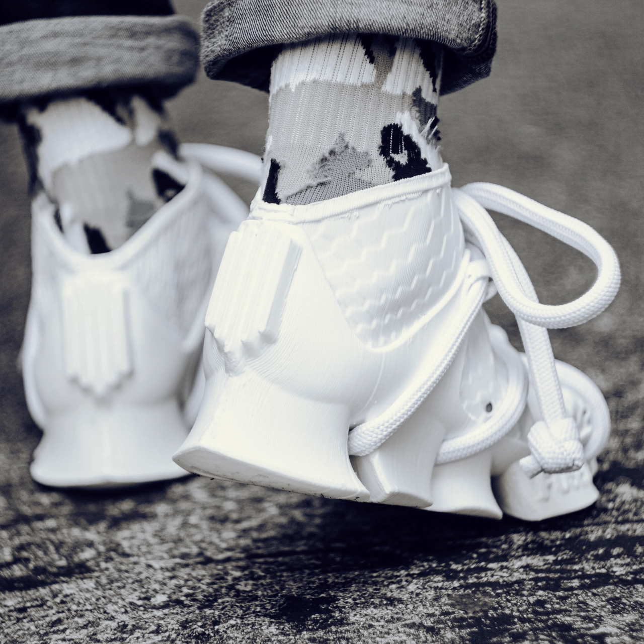 The heels of white rope-laced 3D-printed shoes and patterned socks on asphalt