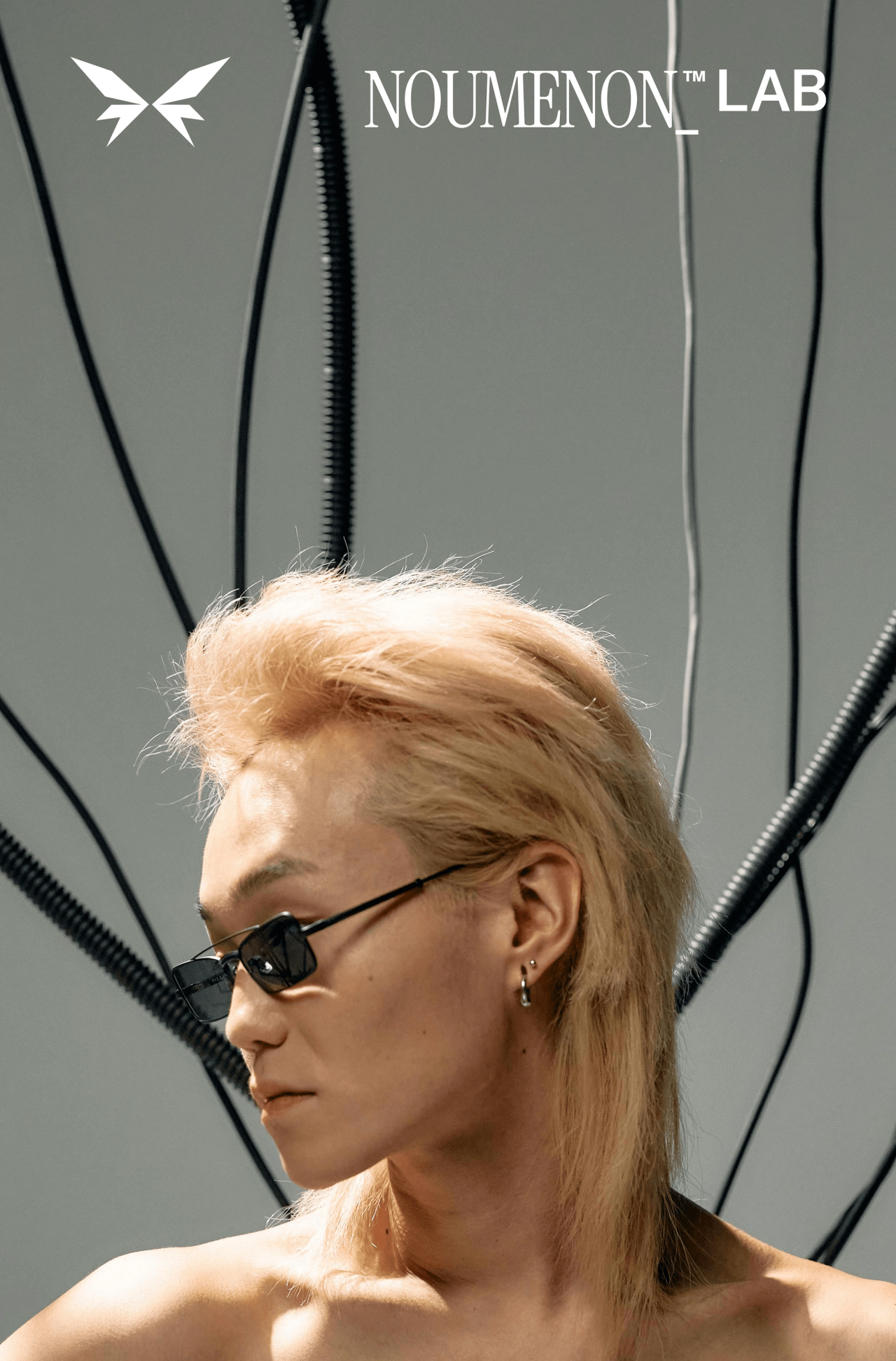 A blond model in narrow black sunglasses among hanging cables, under the NOUMENON LAB logo