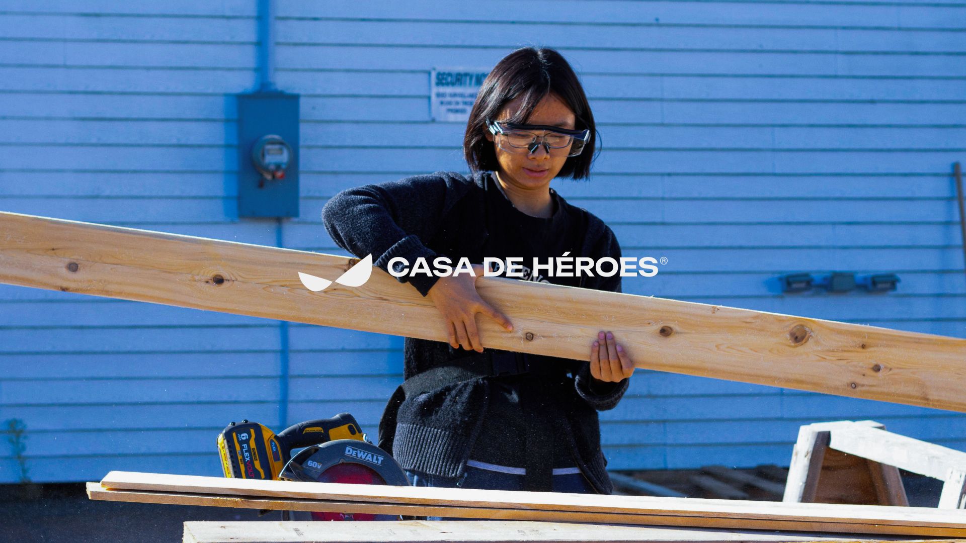 A woman in safety glasses carrying a plank under the Casa de Héroes logo