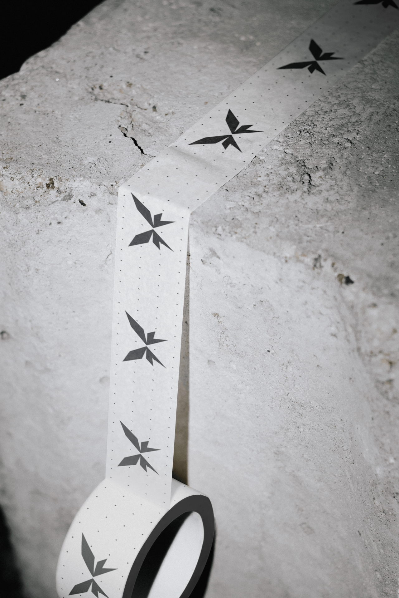 A roll of white tape printed with black NOUMENON LAB butterfly symbols, unrolled across rough stone
