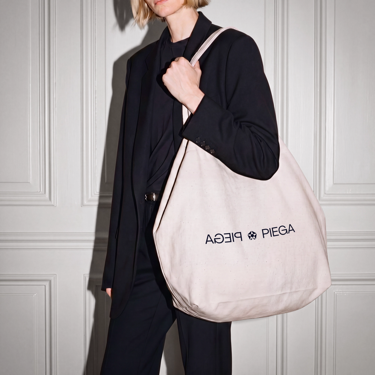 A model in a black suit carrying a canvas tote with the mirrored PIEGA wordmark