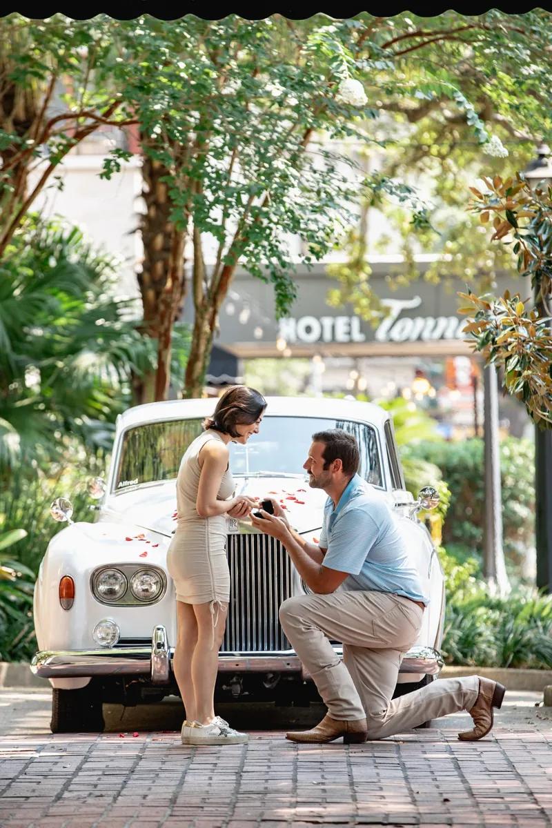 Romantic proposal in New Orleans with man on one knee proposing in front of vintage white Rolls-Royce covered in rose petals.
