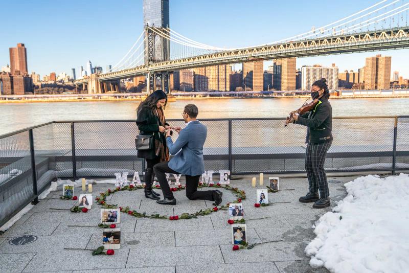 A man is proposing to his partner at a decorated spot on the waterfront in DUMBO, New York. He is kneeling inside a heart made of roses while she smiles and looks at the ring. A violinist stands nearby playing live music. Behind them the Manhattan Bridge and the city skyline stretch across the background, creating a classic New York proposal scene with a romantic atmosphere.