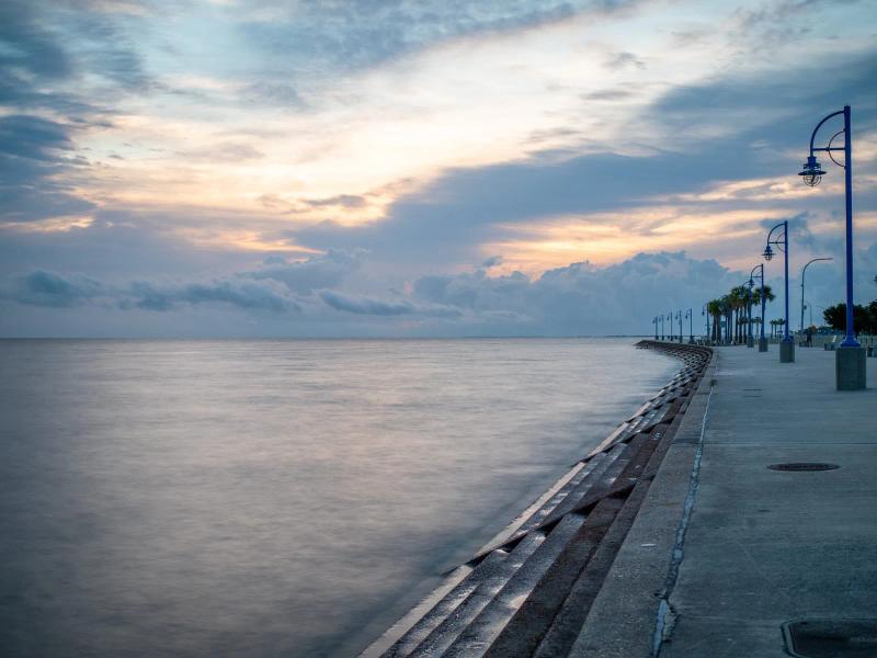 View of the New Orleans Lakefront at sunset, with calm water, a curved concrete walkway lined with blue street lamps, and a colorful sky filled with soft clouds