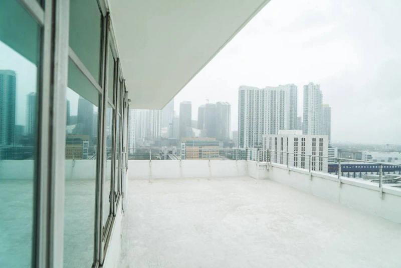 Private rooftop terrace in Miami featuring a spacious white balcony with glass doors and modern railings, overlooking the city skyline of high rise buildings under a soft, overcast sky.