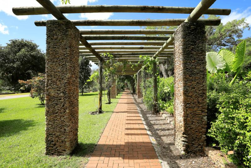 Pergola walkway at Fairchild Tropical Botanic Garden in Miami, featuring stone columns with wooden beams overhead, a brick pathway leading through lush tropical greenery, and bright sunlight casting shadows along the garden path.