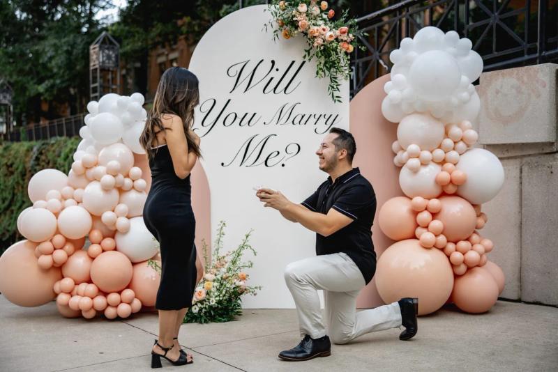 Marriage proposal setup with peach and white balloon arch and “Will You Marry Me?” sign, man on one knee proposing to woman in black dress.